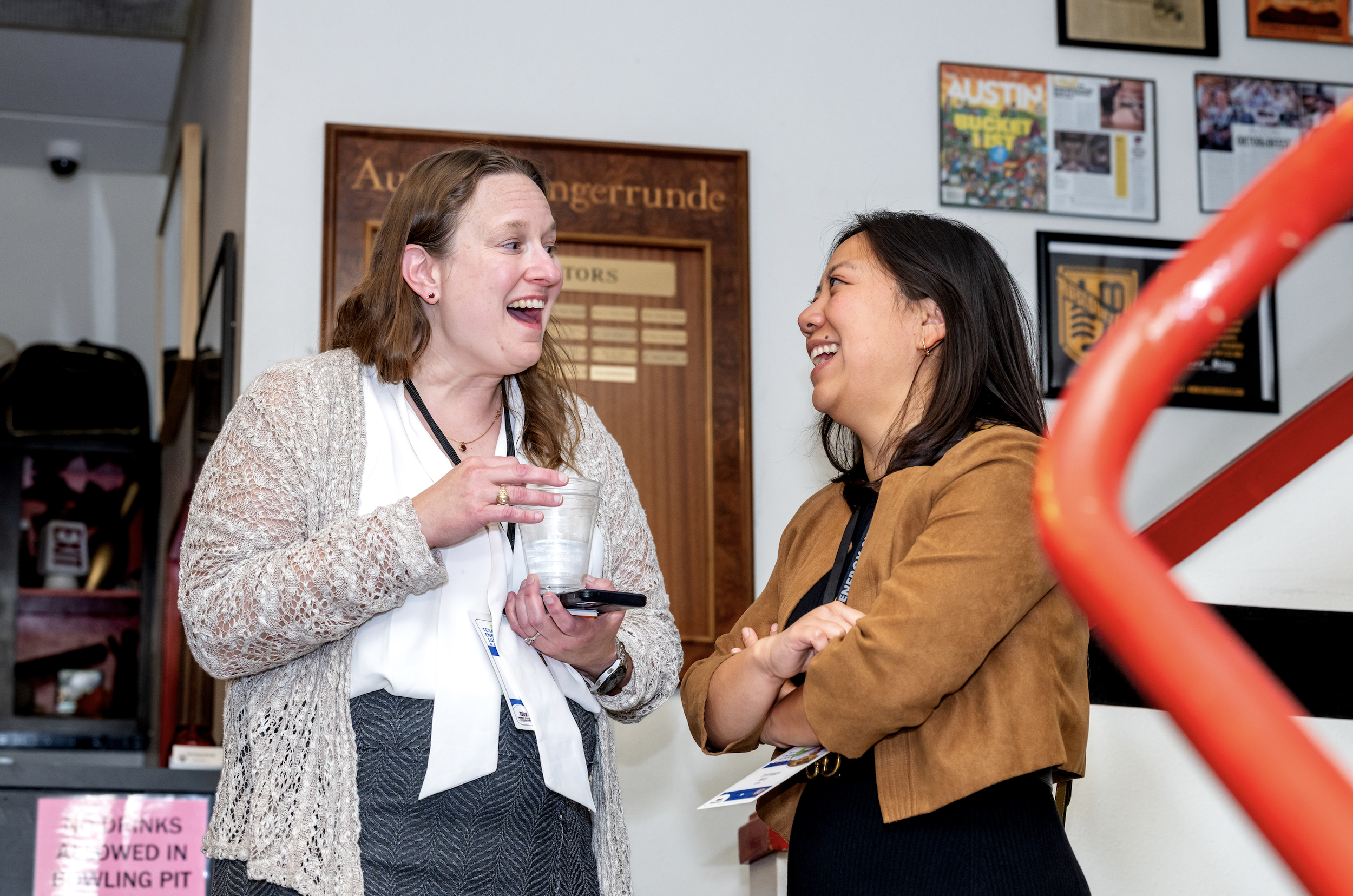 Two women smile and laugh while talking indoors, one holding a glass and a phone, in a room decorated with framed pictures and a wooden plaque.
