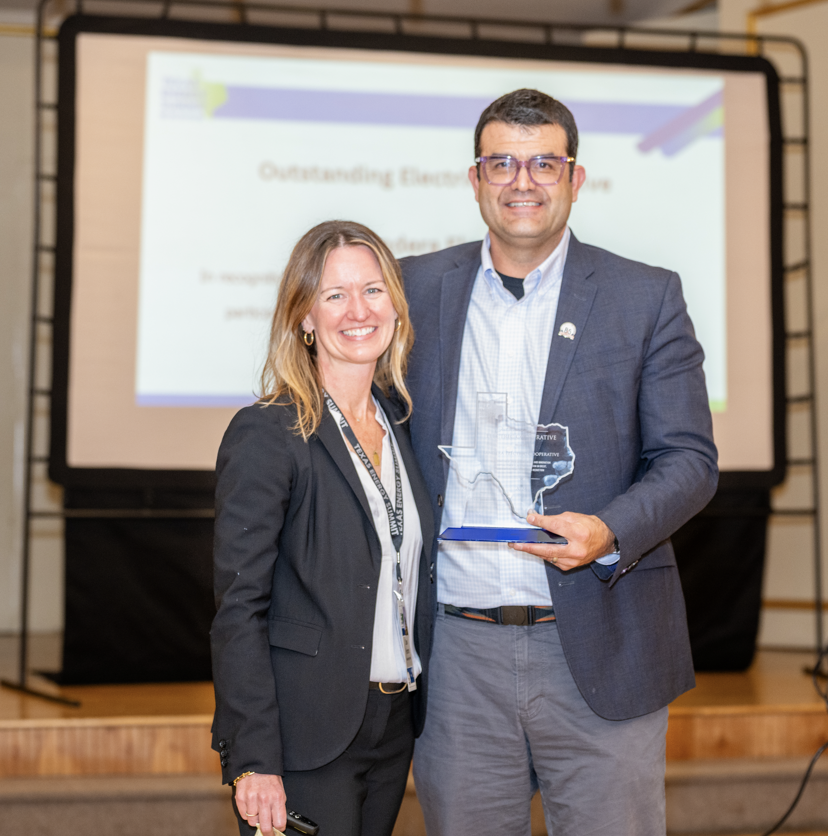 A woman and a man are smiling and standing side by side, holding a clear award shaped like Texas. They are at a formal event with a presentation screen in the background displaying the words 'Outstanding Element...'