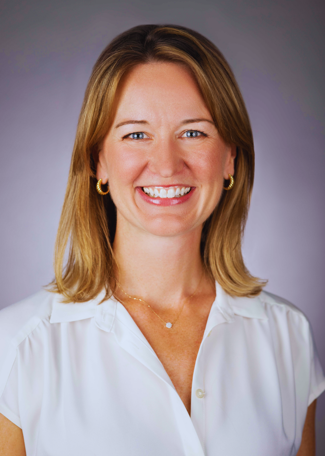 A woman with shoulder-length blonde hair, blue eyes, and a bright smile, wearing gold hoop earrings, a delicate necklace, and a white blouse, against a gray background.