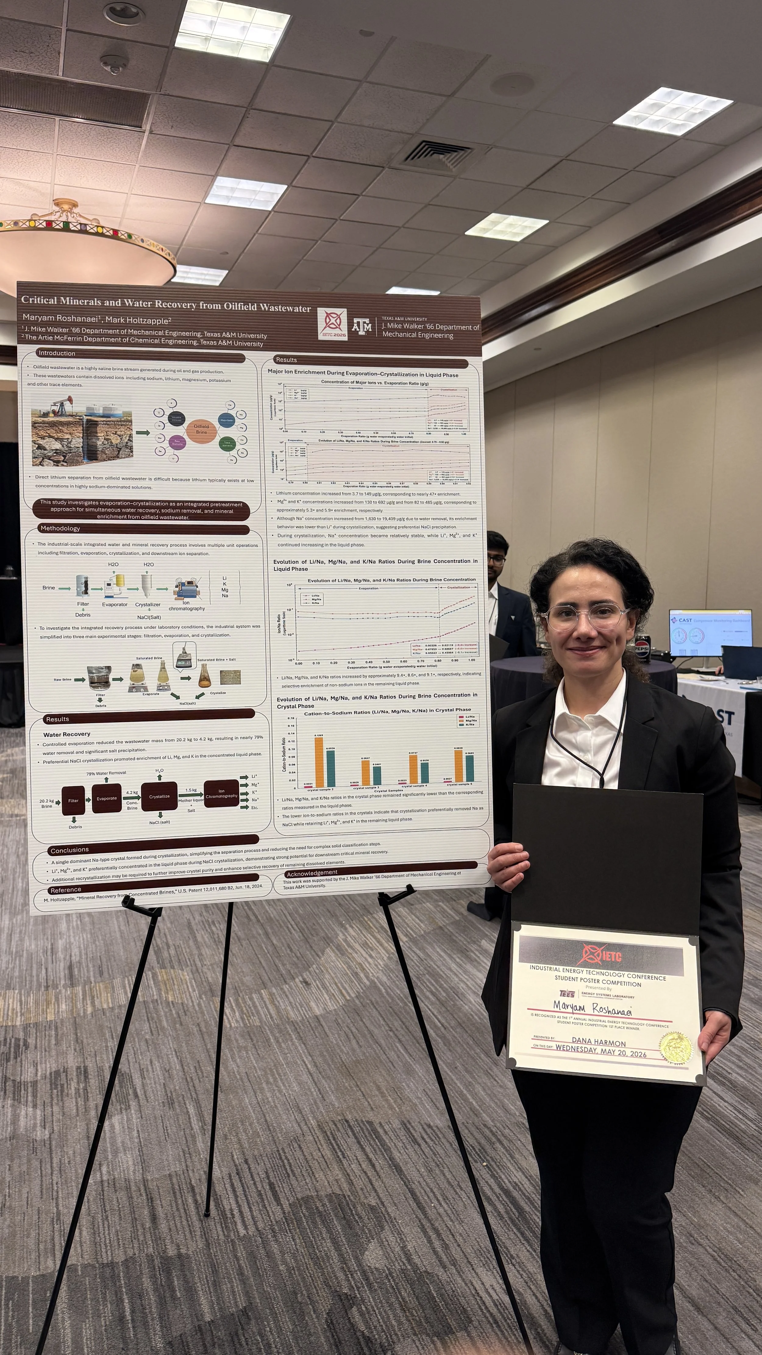 A woman standing next to a research poster at a conference, wearing glasses, a light-colored blazer, matching pants, and white shoes.