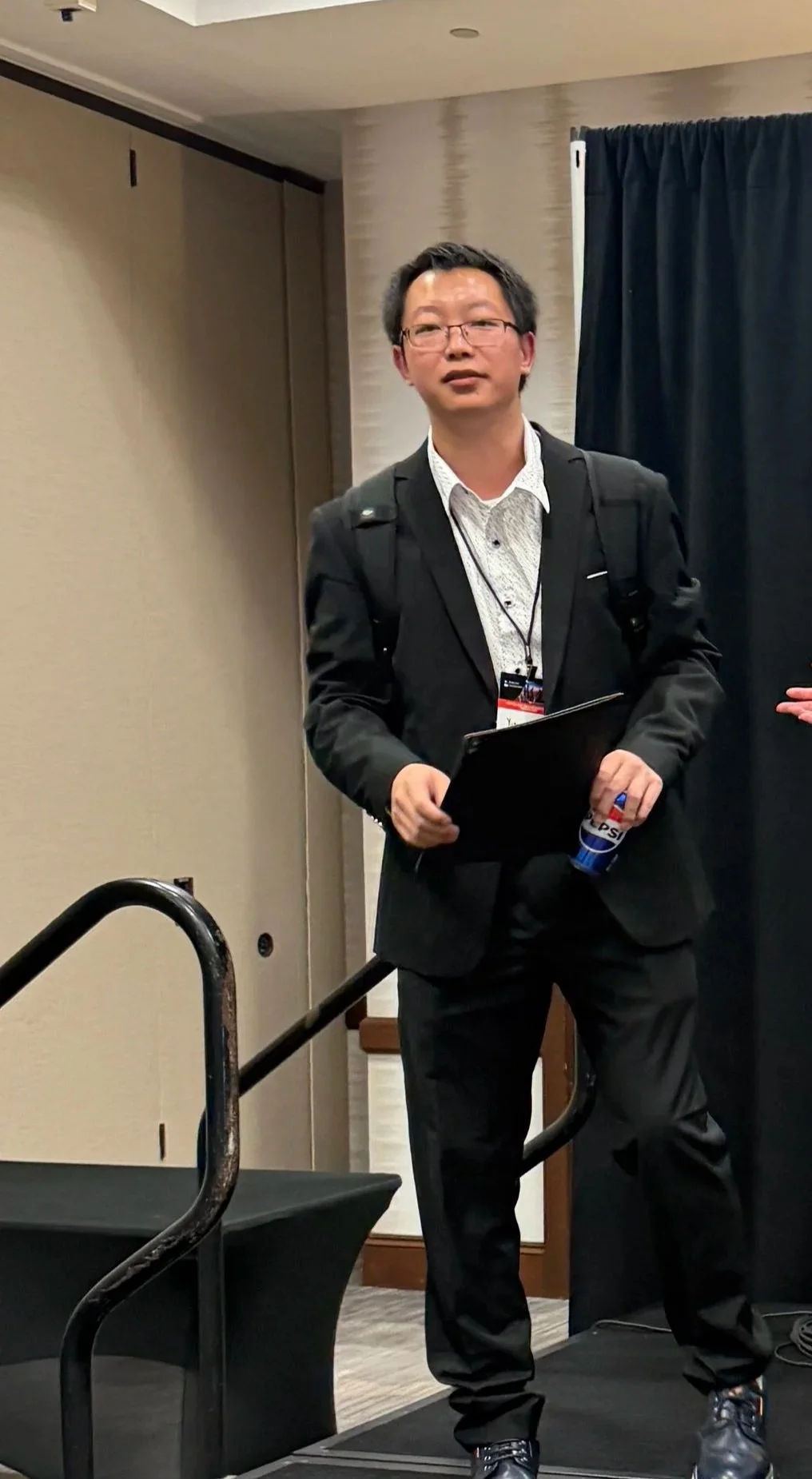 A man in formal attire standing next to a scientific research poster at a conference. The poster discusses managing the impact of electric vehicles on utility grids, with sections on introduction, existing solutions, case-study community, energy efficiency programs, and future work.