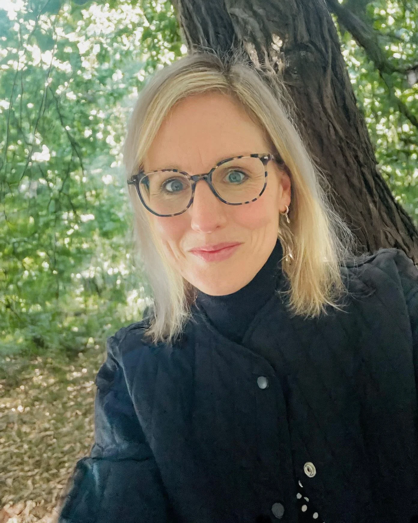A woman with blonde hair, glasses, and earrings taking a selfie outdoors under a tree with green leaves.