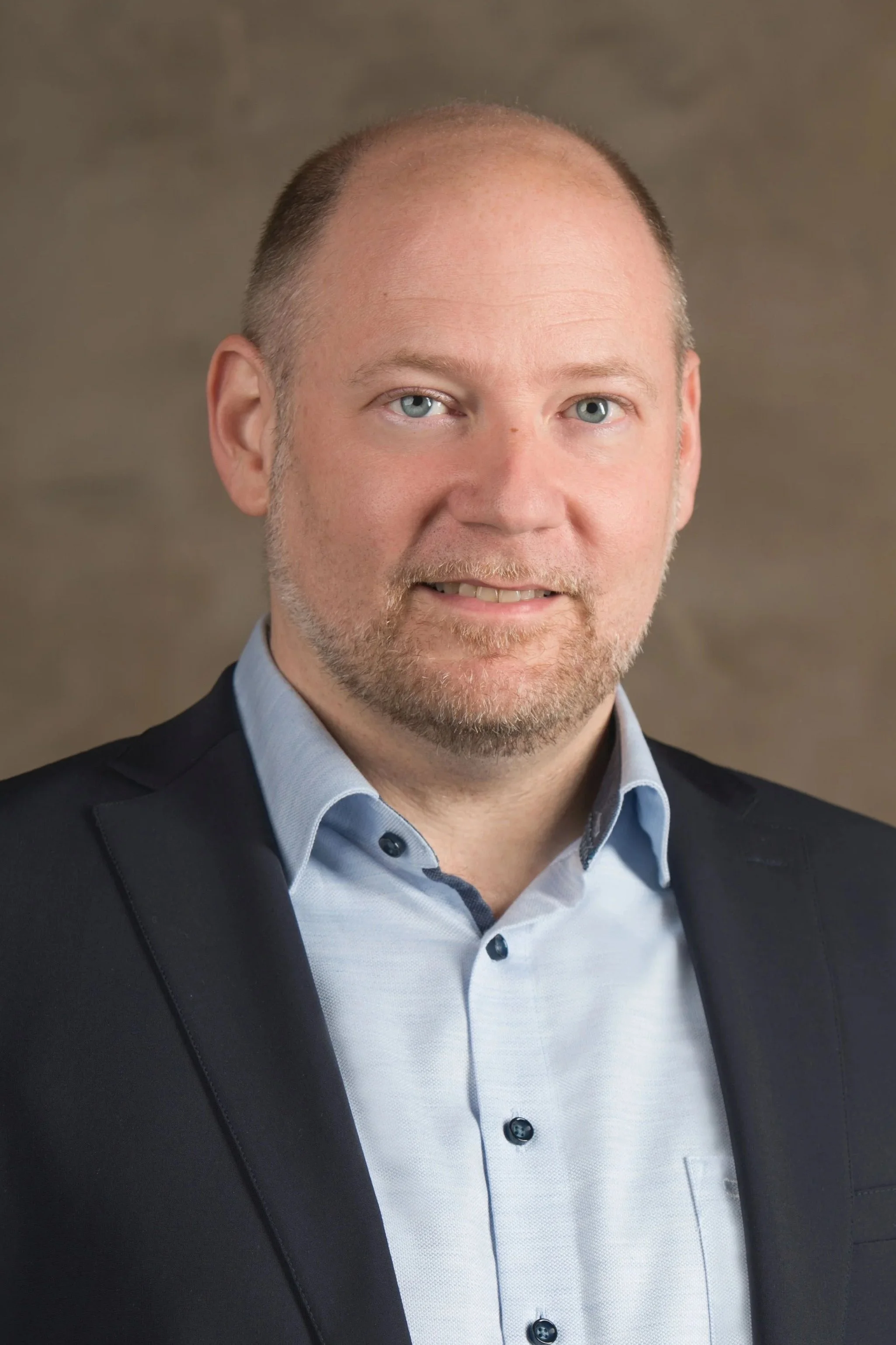Porträt eines Mannes in einem blauen Hemd und dunklem Blazer vor braunem Hintergrund.
