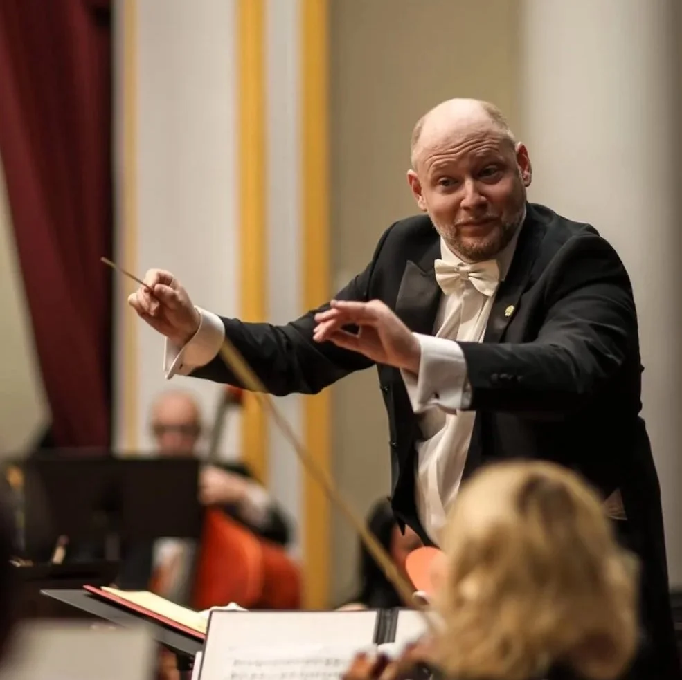 Ein Dirigent leitet ein Orchester während einer Aufführung, trägt einen schwarzen Frack und eine weiße Fliege, mit Orchesterstücken im Hintergrund.