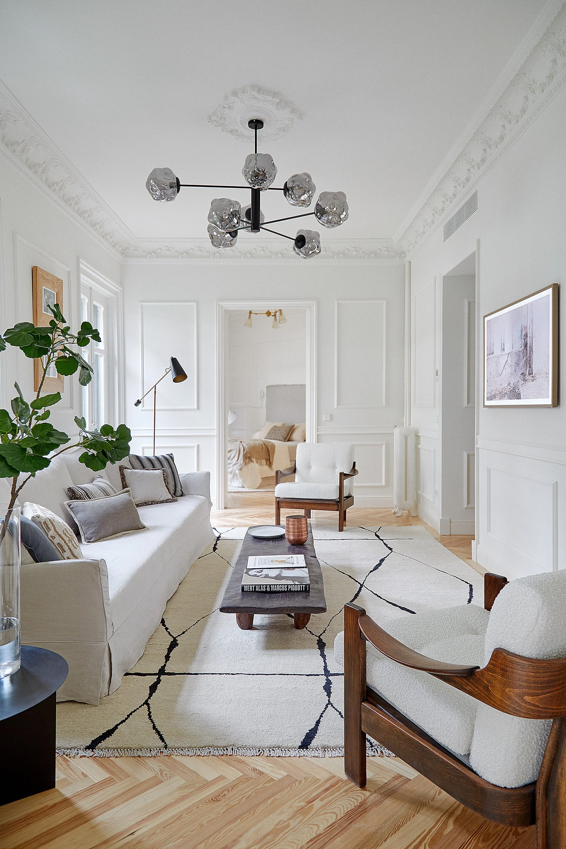 Sala de estar con sillones blancos, una mesa de madera oscura, una alfombra blanca con patrón negro, plantas verdes, y paredes blancas con molduras decorativas.