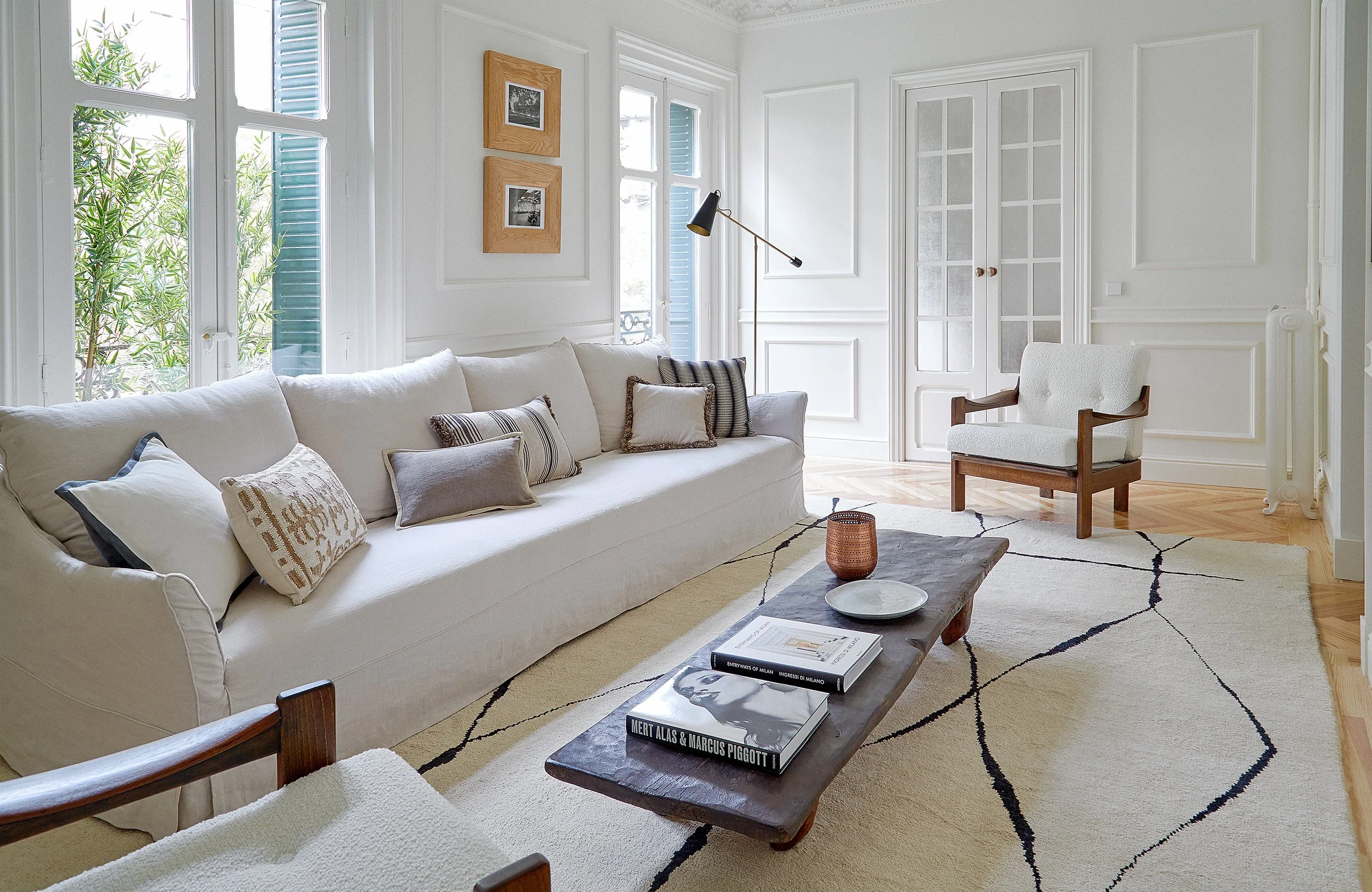 Sala de estar con sofá blanco, varias almohadas y mesa de madera con libros y objetos decorativos, ventanas con cortinas, pared blanca y piso de madera