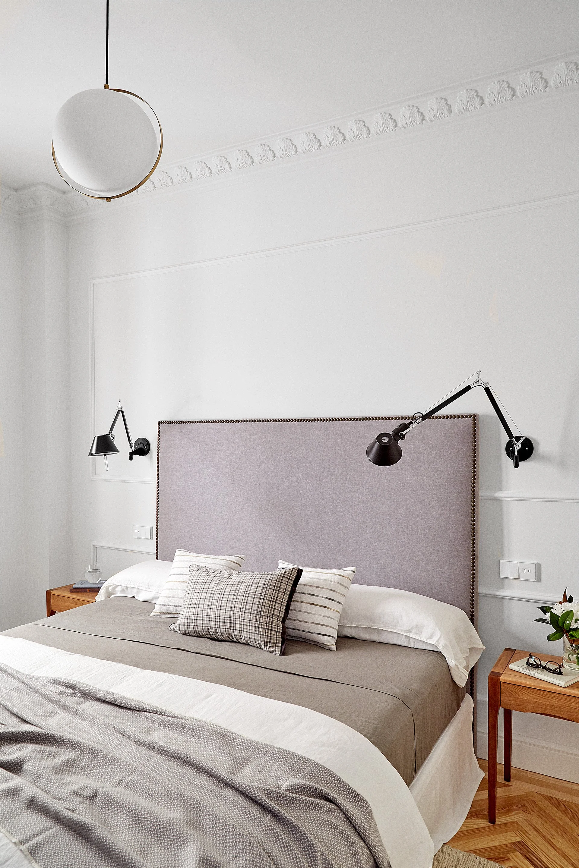 Dormitorio con cabecera de tela gris, paredes blancas, lámparas ajustables negras en ambos lados, y mesas de madera con objetos decorativos y plantas.