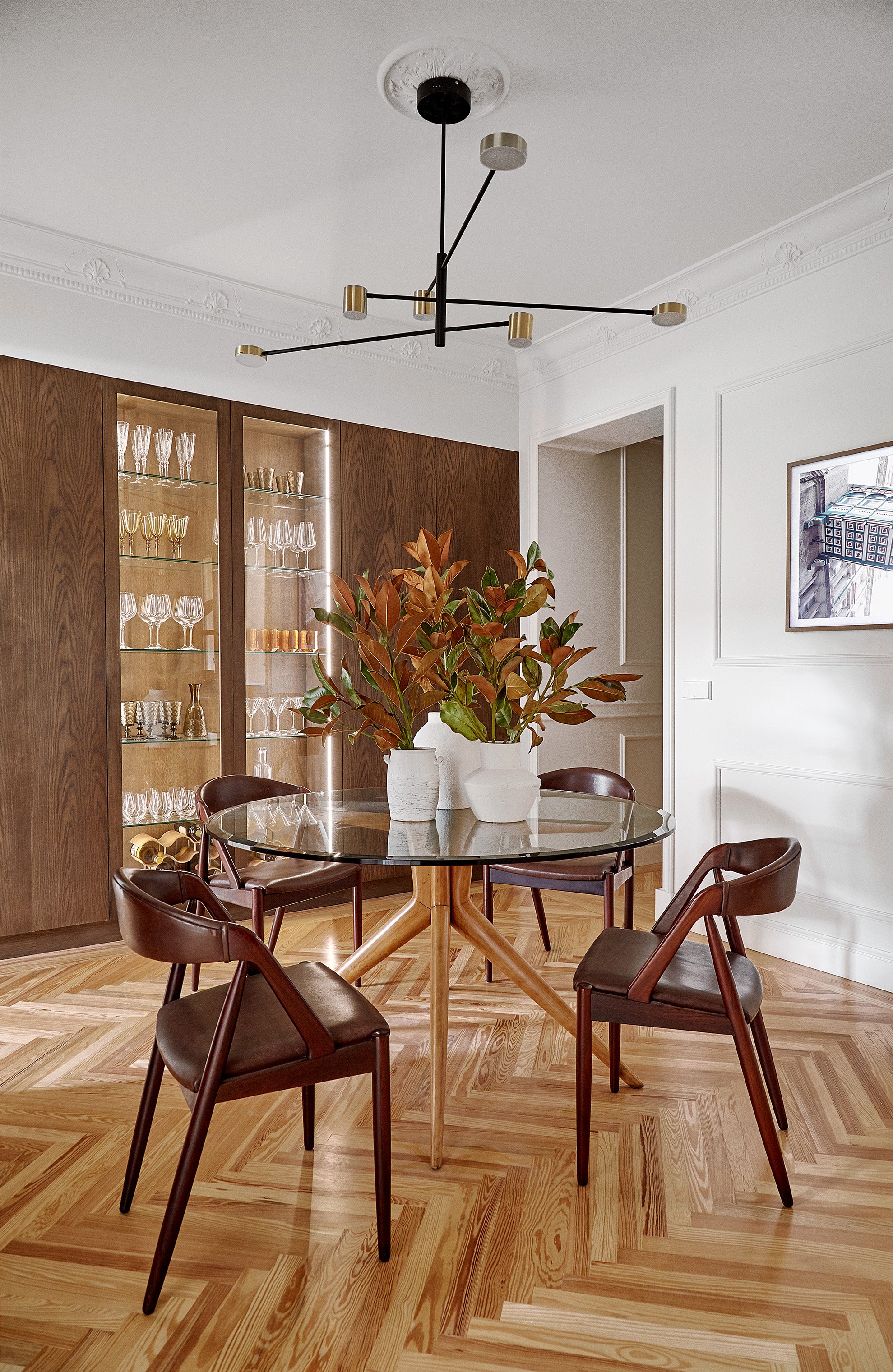 Comedor con mesa redonda de vidrio, cinco sillas de madera con asientos de cuero, una planta en un florero blanco en el centro y un mueble de madera con cristalería en el fondo. Techo con lámpara moderna y piso de madera.