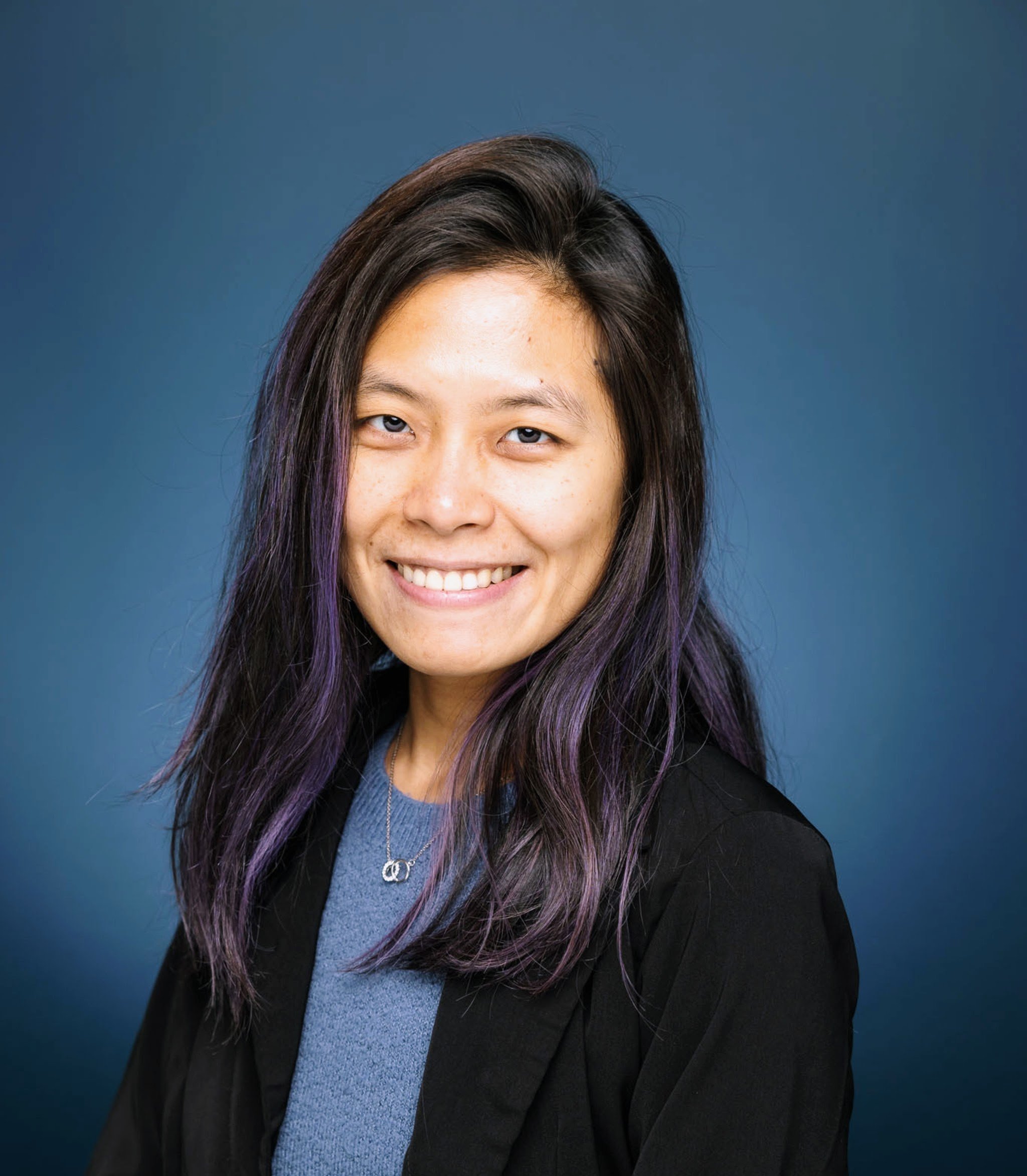 Smiling woman with long dark hair with purple streaks, wearing a black blazer over a blue top, against a blue background.