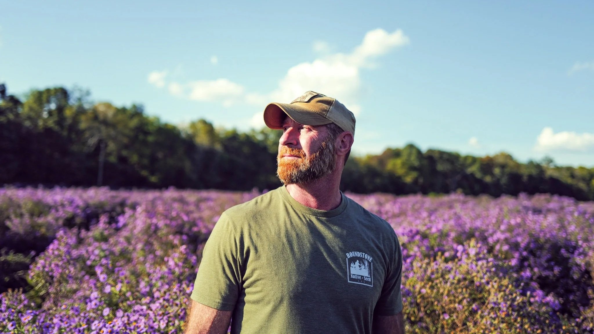 Man with a beard and cap standing in a field of purple flowers under a blue sky.