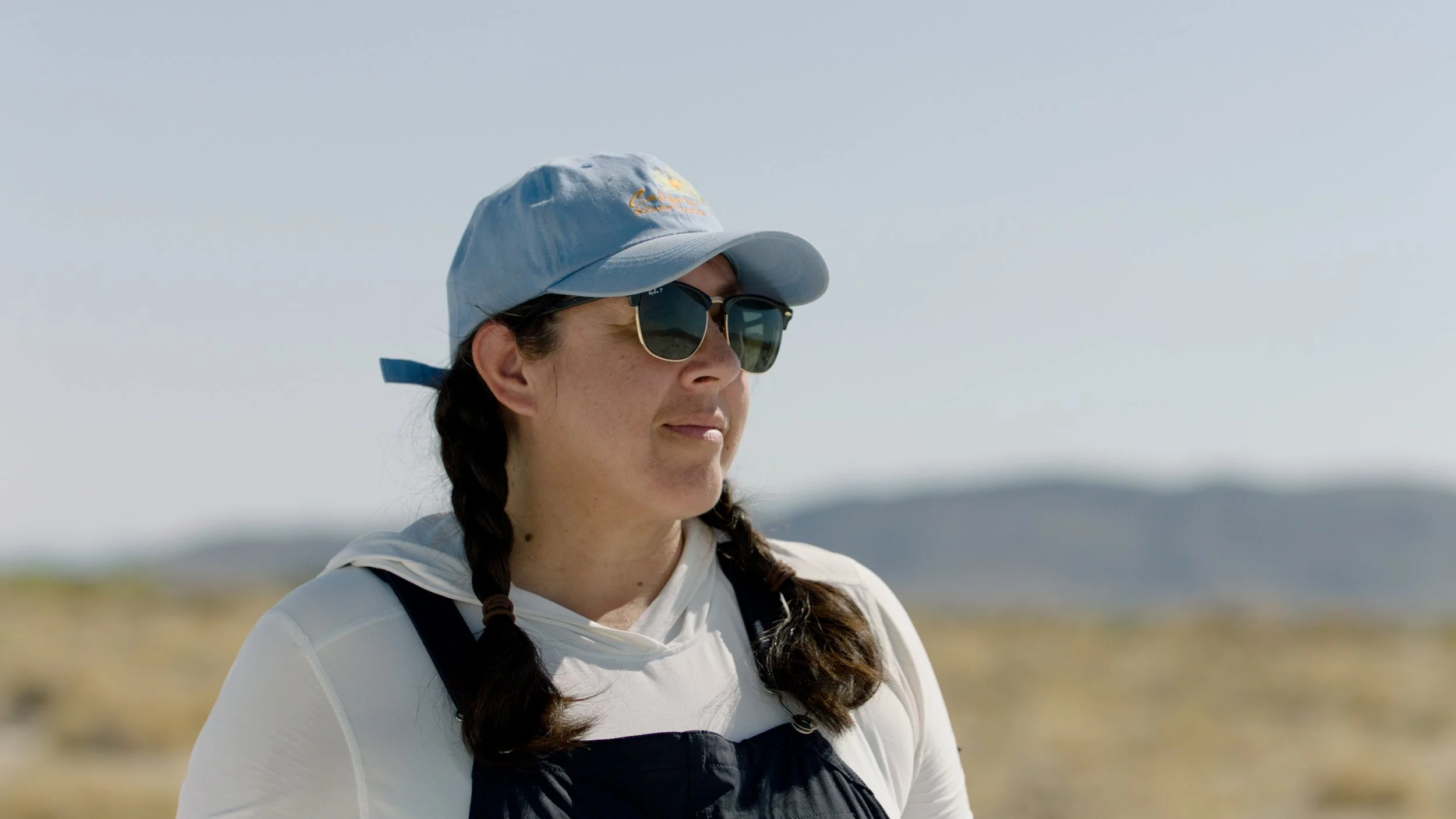 A woman wearing sunglasses, a light blue cap, and a white hoodie outdoors with a blurred landscape of mountains and sky in the background.