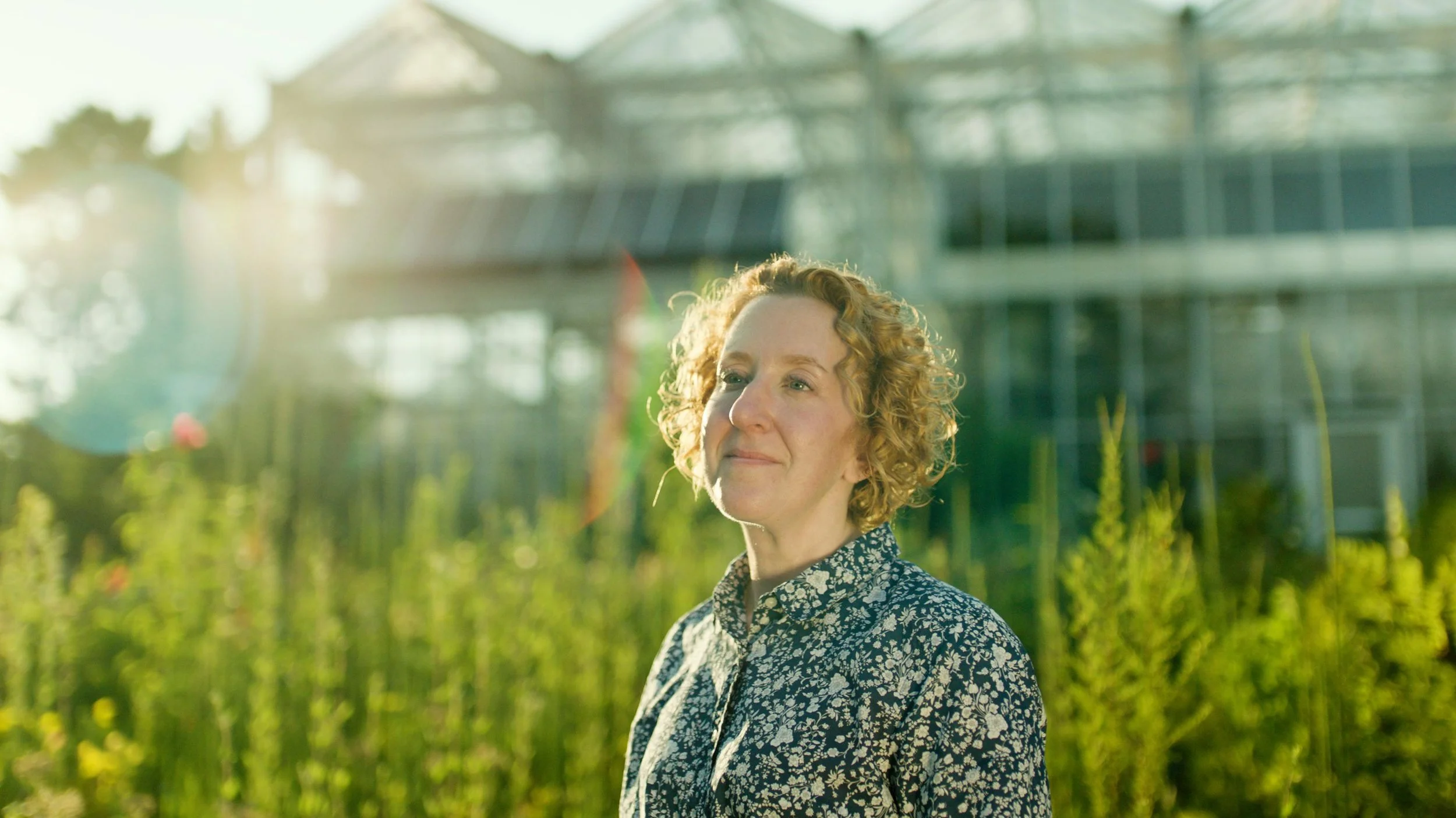A woman with curly red hair standing outdoors in a greenhouse garden during daylight, wearing a floral patterned shirt, with sunlight shining and greenery in the background.