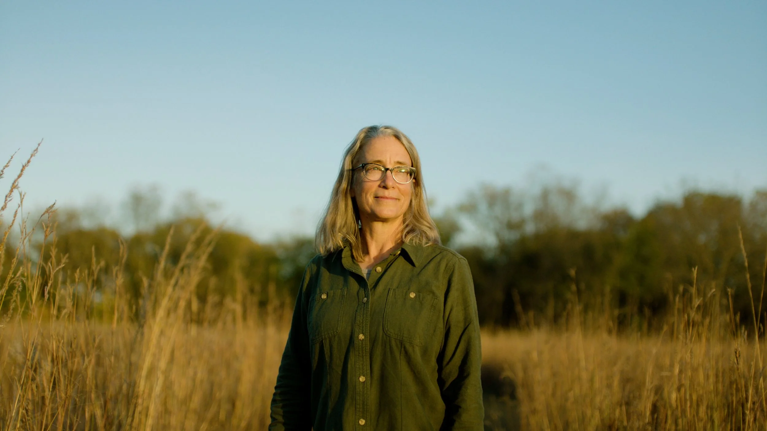 A woman with glasses and shoulder-length blonde hair standing in a field of tall grass, looking into the distance, with trees and a clear blue sky in the background, during daytime.