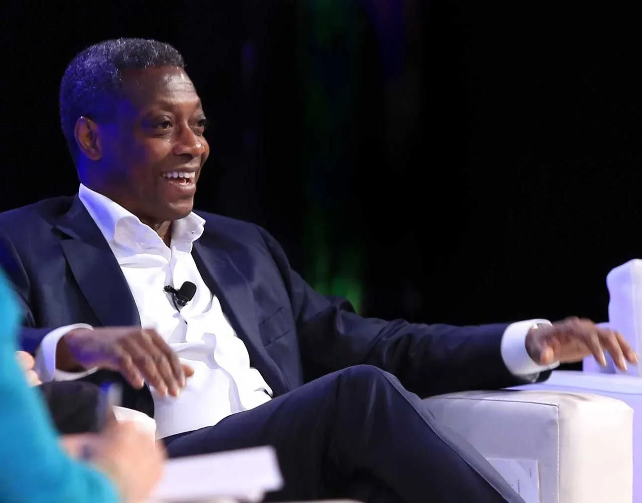 A man in a formal suit sitting on a white chair, smiling and talking during a discussion or interview, with a dark background.