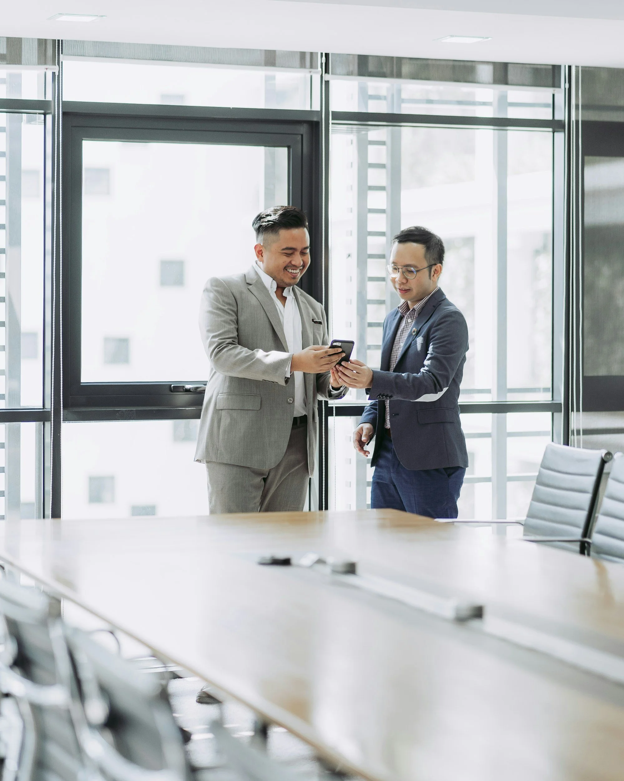 Two business professionals in suits standing in a modern office, sharing a laugh while looking at a smartphone, with a large conference table and office windows in the background.