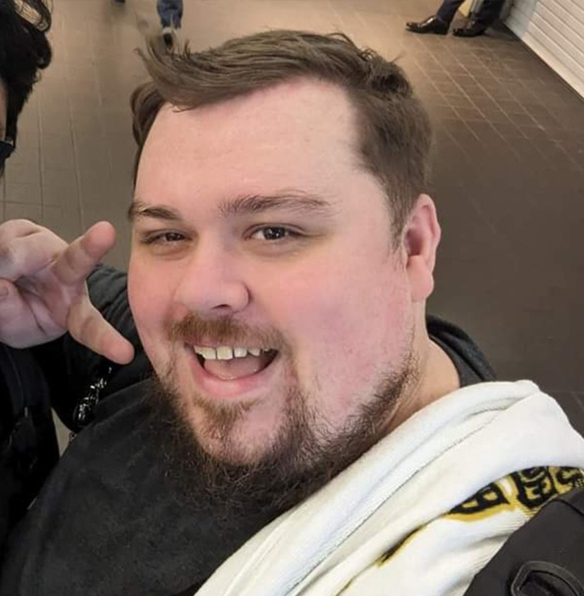 A smiling man with a beard and mustache taking a selfie, making a hand gesture, in an indoor setting with people in the background.