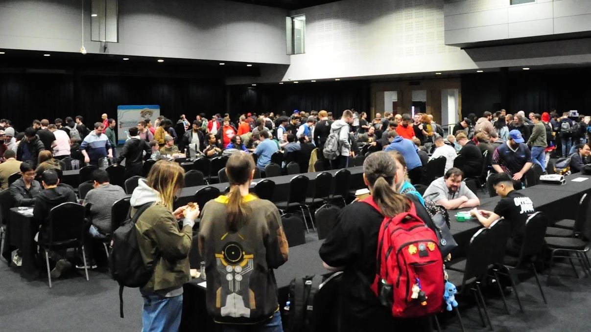 Large indoor event with many people sitting and standing at tables, some engaged in card games, in a conference hall or convention center.