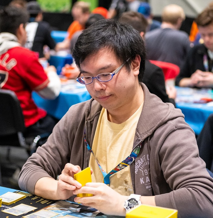A man with glasses in a brown hoodie playing a card game at a table during a crowded event.