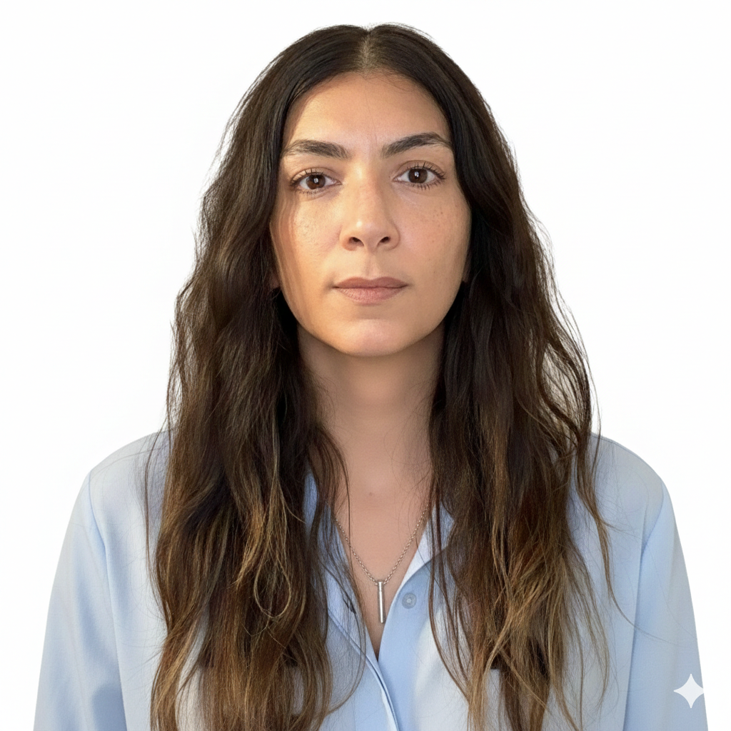 A woman with long wavy brown hair, wearing a light blue blouse, and a silver necklace, facing the camera against a white background.
