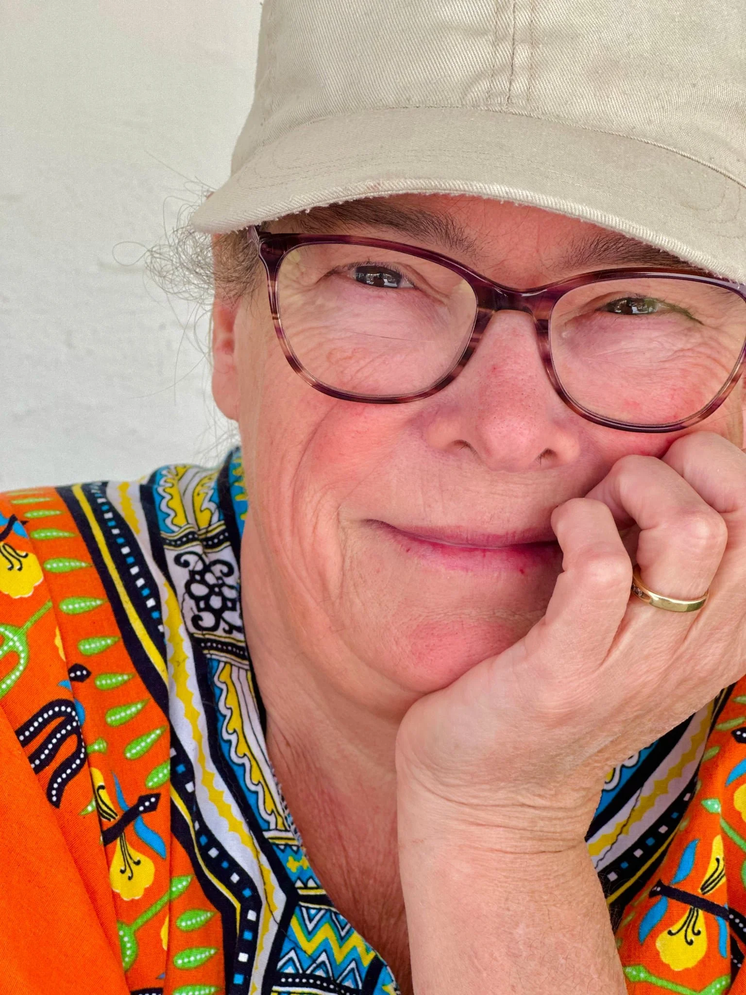 A woman wearing glasses, a beige hat, and a bright colorful garment, resting her face on her hand and smiling gently.