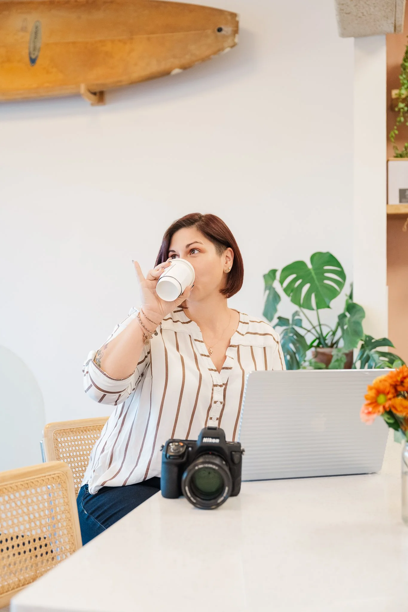 A woman with short brown hair and a striped shirt sitting at a table, drinking from a paper cup, with a camera and a laptop in front of her, in a room decorated with plants and a wooden surfboard on the wall.