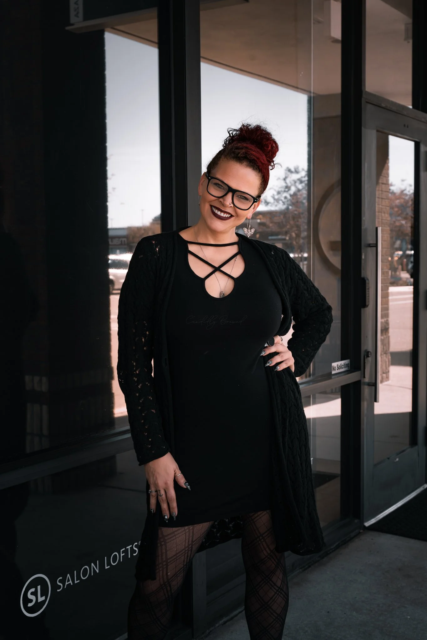 A woman with glasses and red hair styled in a bun, wearing a black dress with crisscross straps, lace cardigan, and fishnet stockings, standing outside a glass door with a sign that reads 'Salon Lofts'.