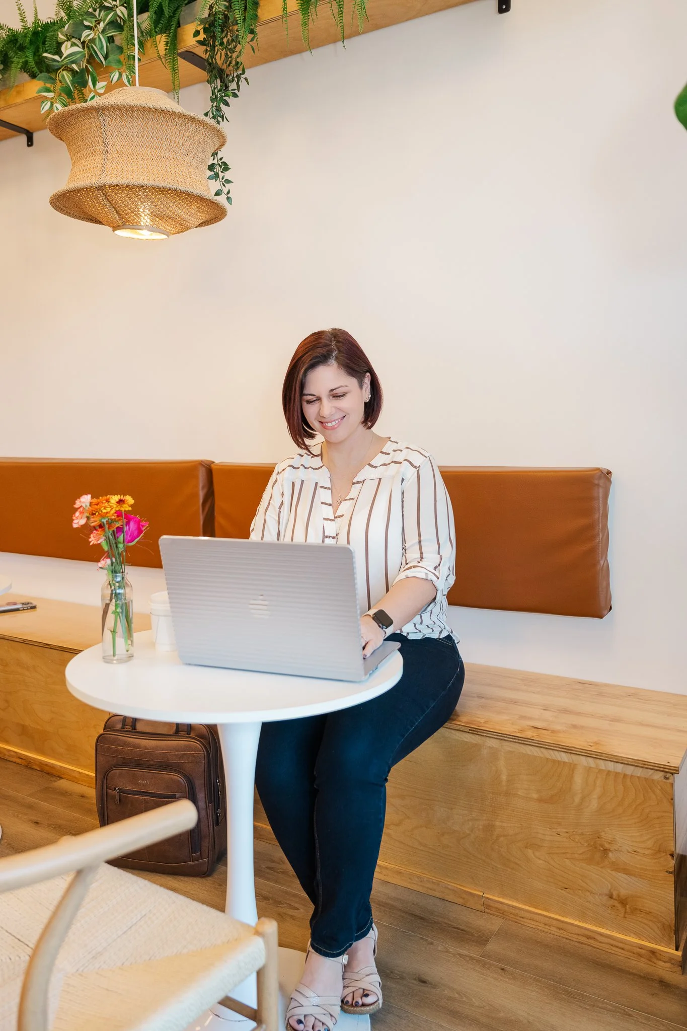 A woman sitting at a white round table with a laptop, a small vase of flowers, a coffee cup, and a backpack underneath. She is smiling and in a cafe or coffee shop with wooden accents and indoor plants.