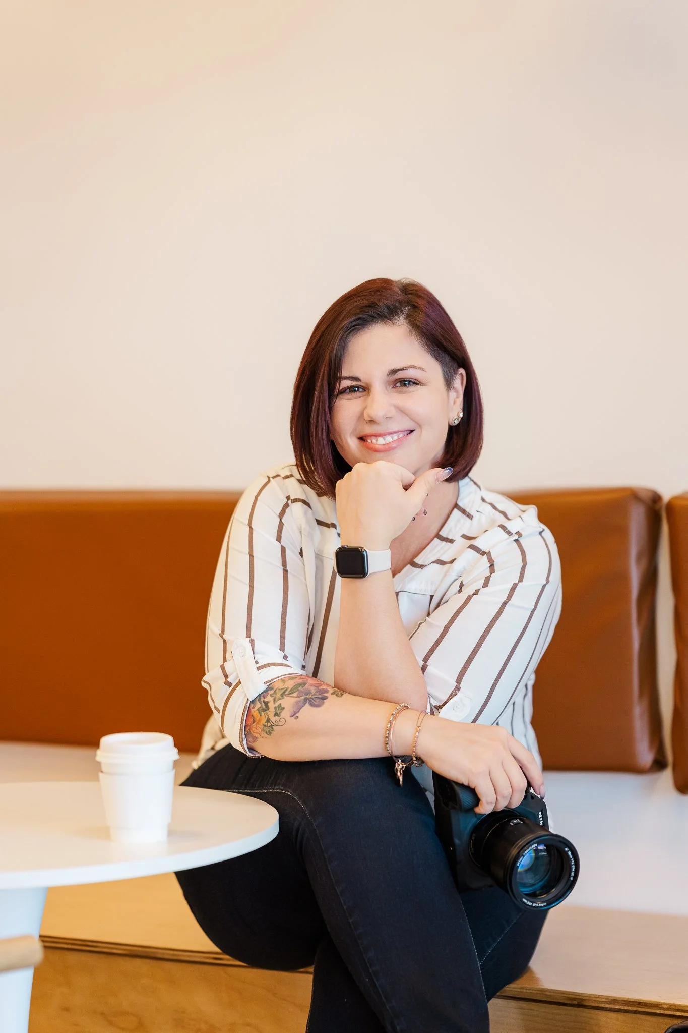 A smiling woman with short dark red hair, wearing a striped shirt, sitting on a bench with a camera in her lap and a smartwatch on her wrist, in a cozy indoor setting.