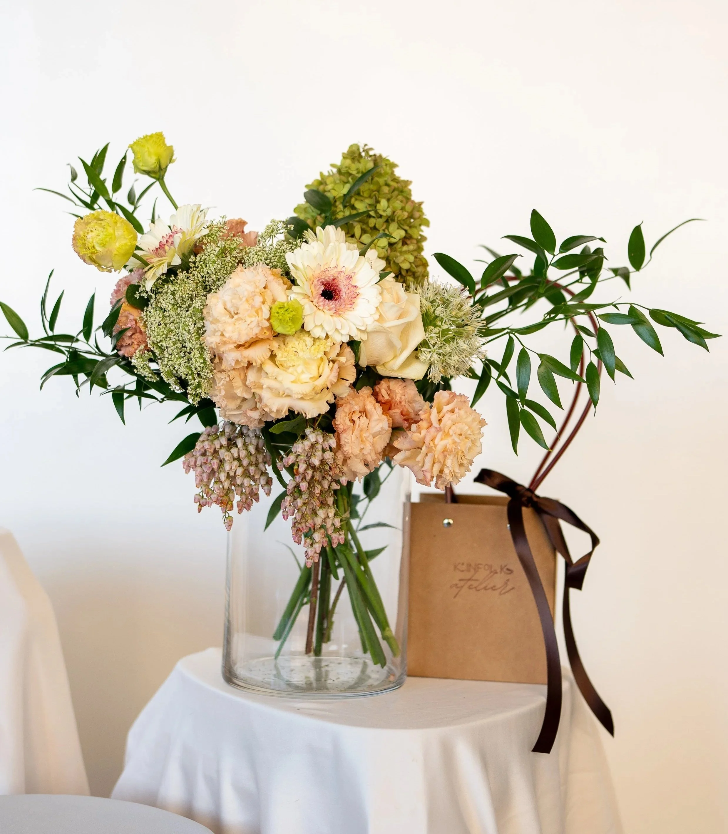 A bouquet of pastel-colored flowers in a glass vase on a white table, with a small brown gift bag behind it.