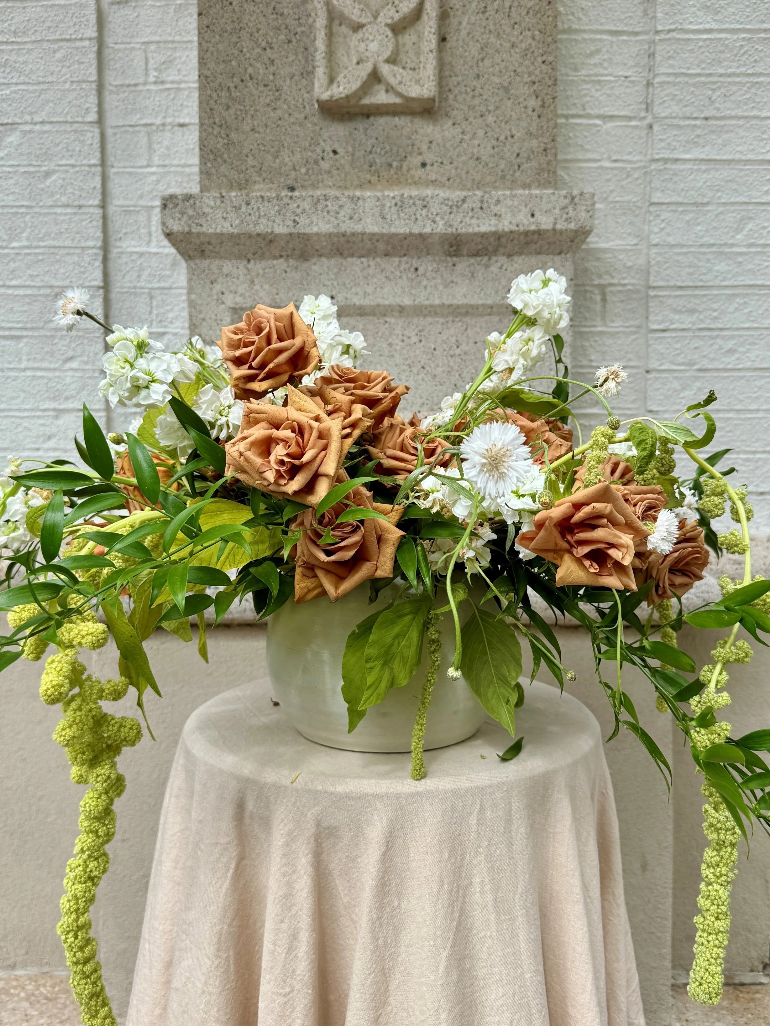 A floral arrangement with green, brown and white flowers in a white vase, placed on a beige tablecloth-covered table against a stone and brick wall background.