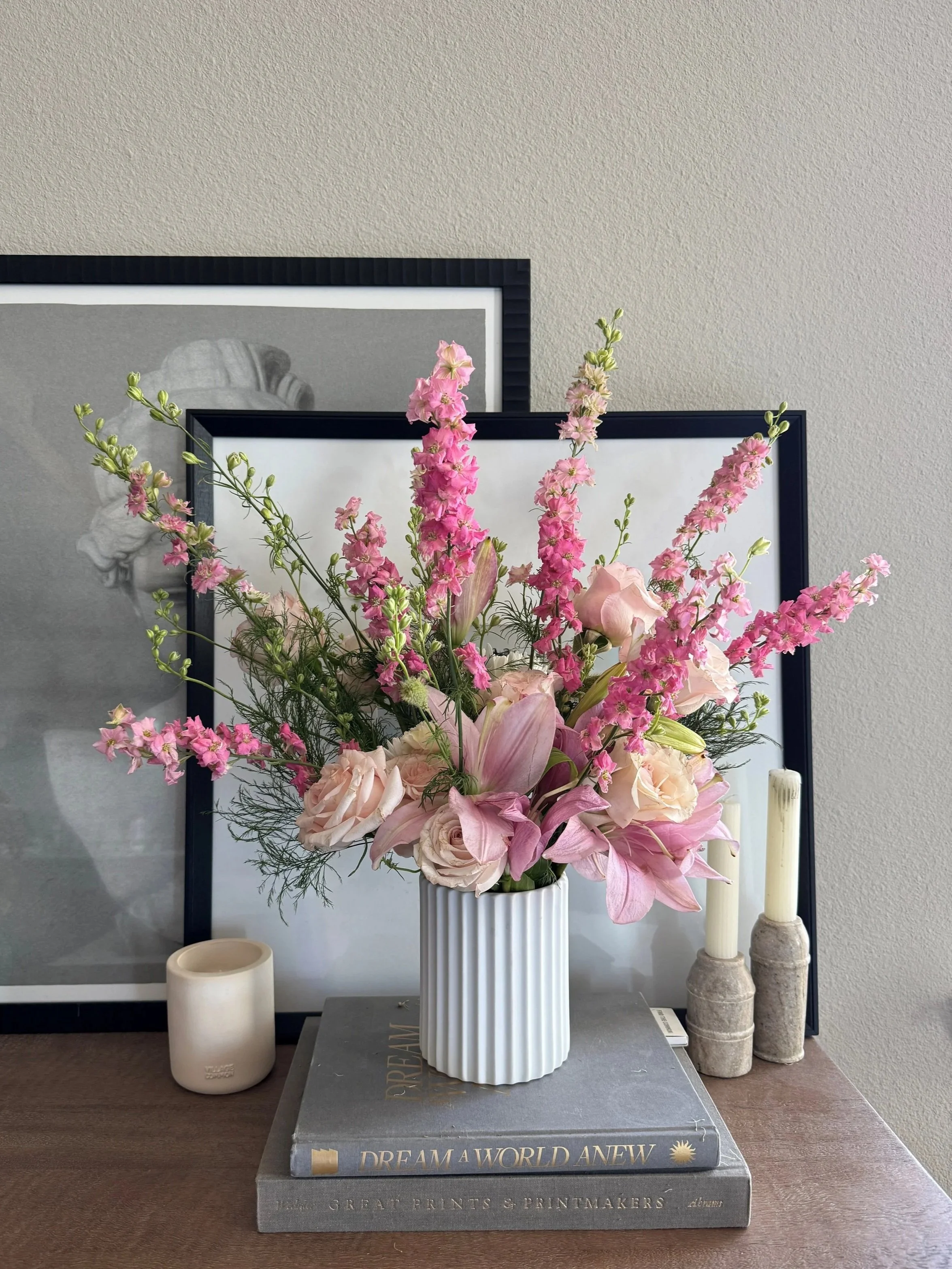 A floral arrangement with pink and white flowers in a white vase, placed on top of two books, with framed monochrome pictures and candles in the background.