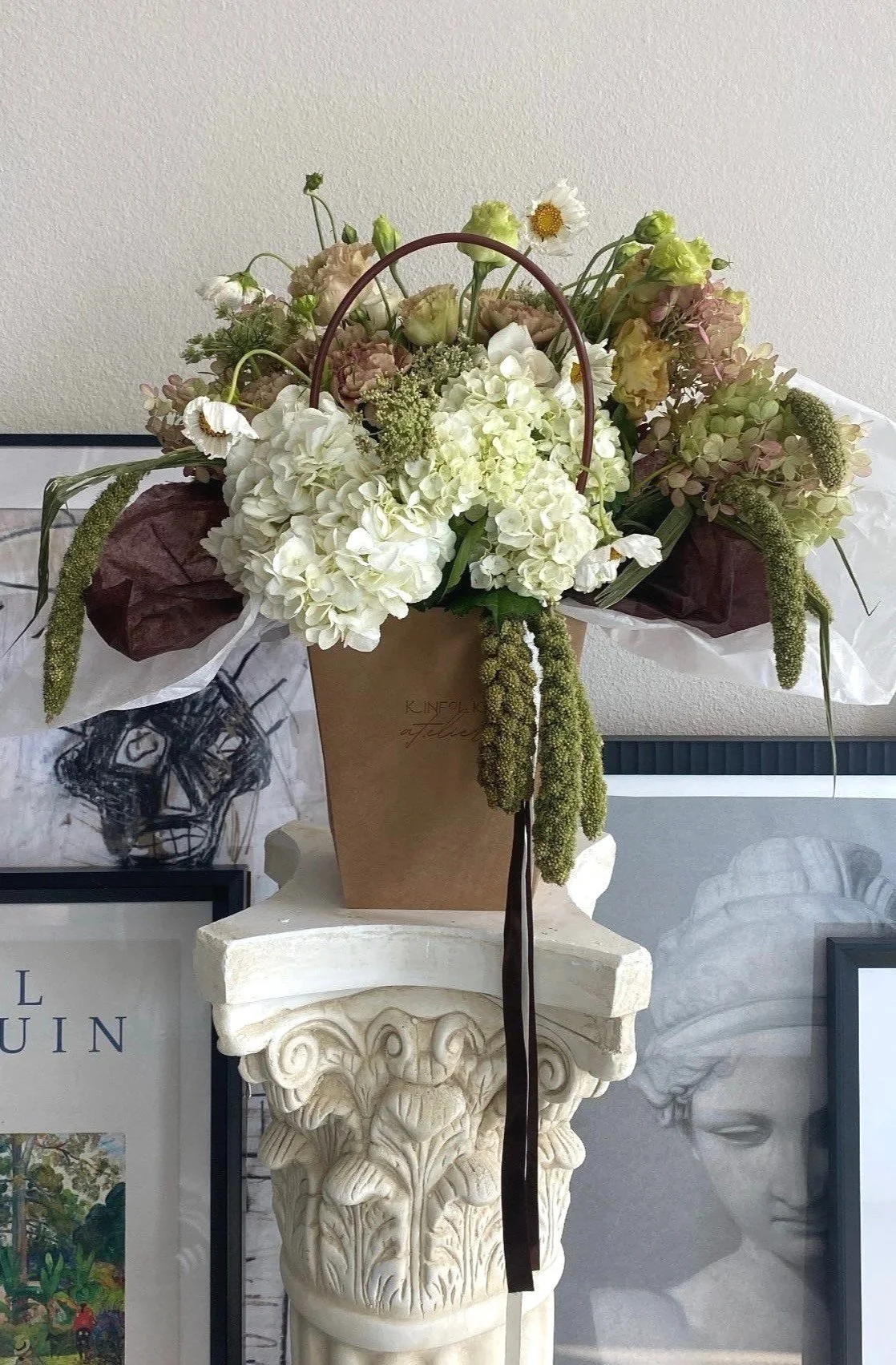 A bouquet of white and pastel-colored flowers in a paper bag placed on a decorative stone pedestal, with framed artwork in the background.