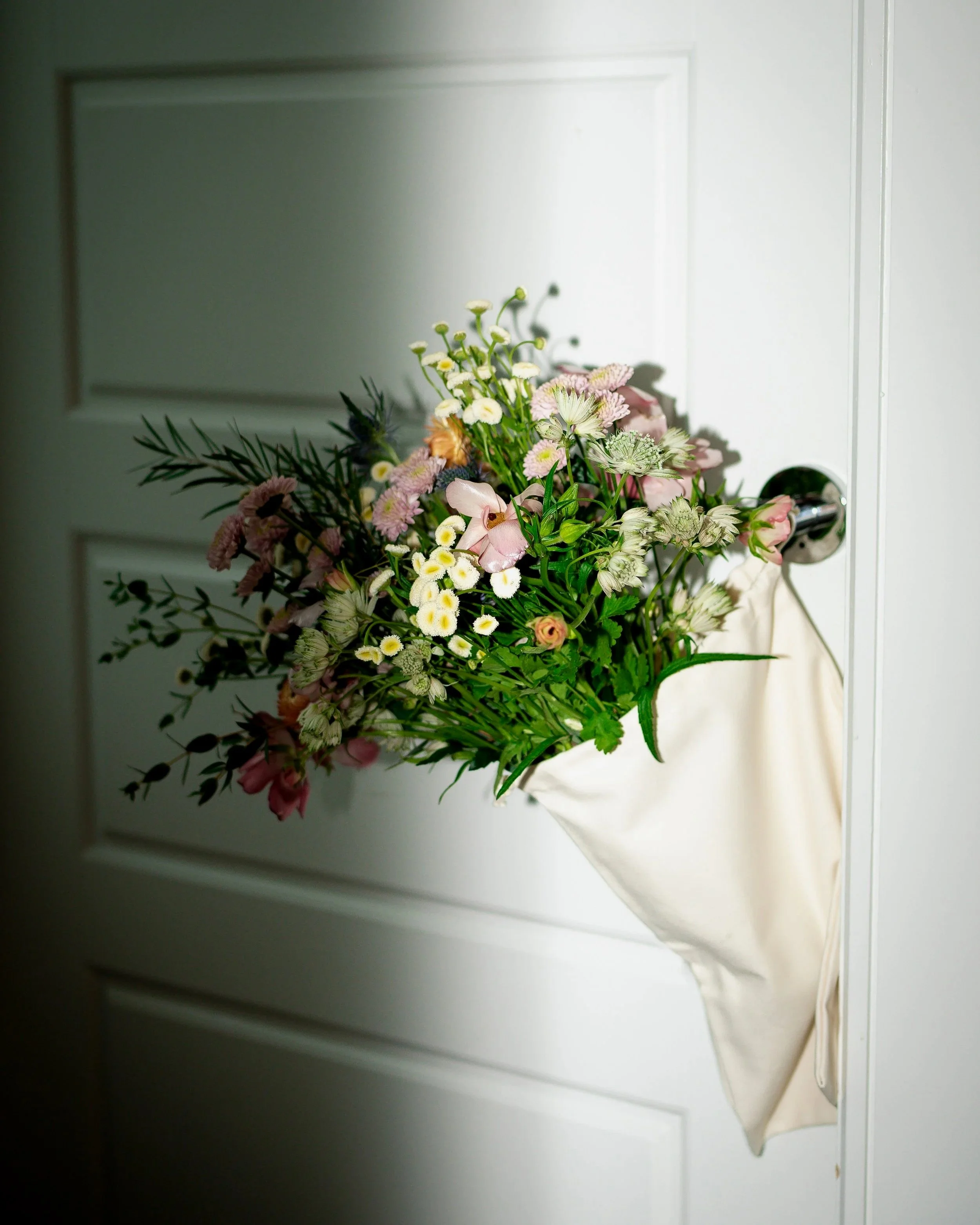 A bouquet of mixed flowers hanging on a door handle with a white canvas bag.