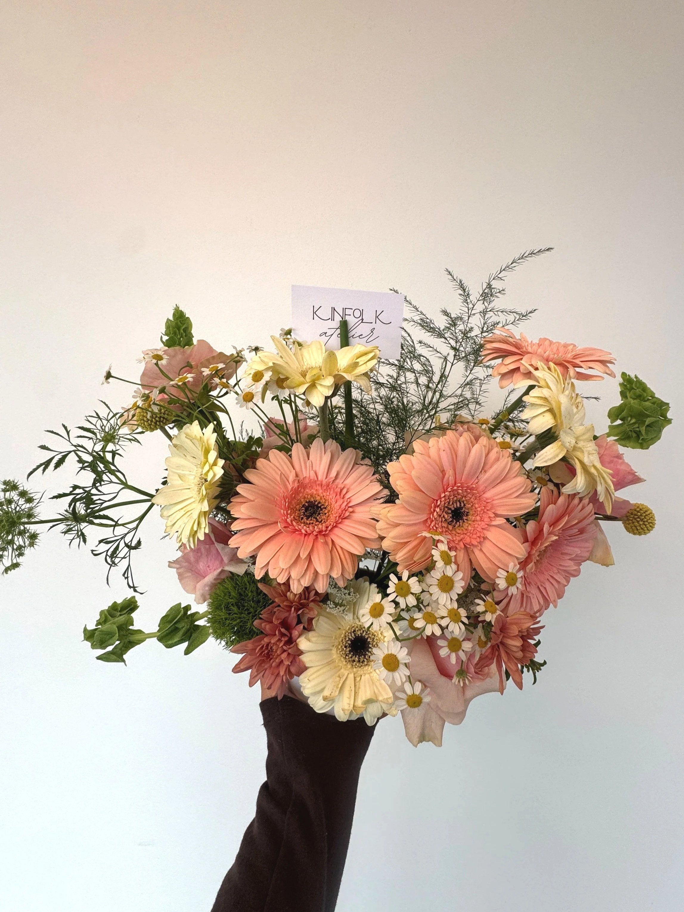 A person holding a bouquet of mixed pastel-colored flowers, including daisies, gerbera daisies, and lilies, against a plain white wall background.
