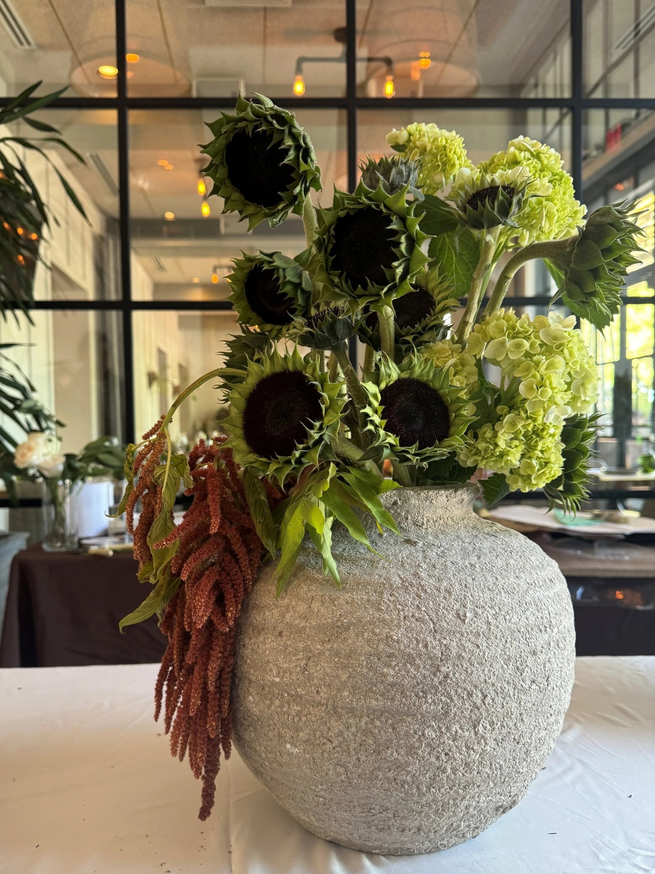 A large textured gray vase filled with sunflowers, hydrangeas, and other greenery, placed on a white table in a modern indoor setting.