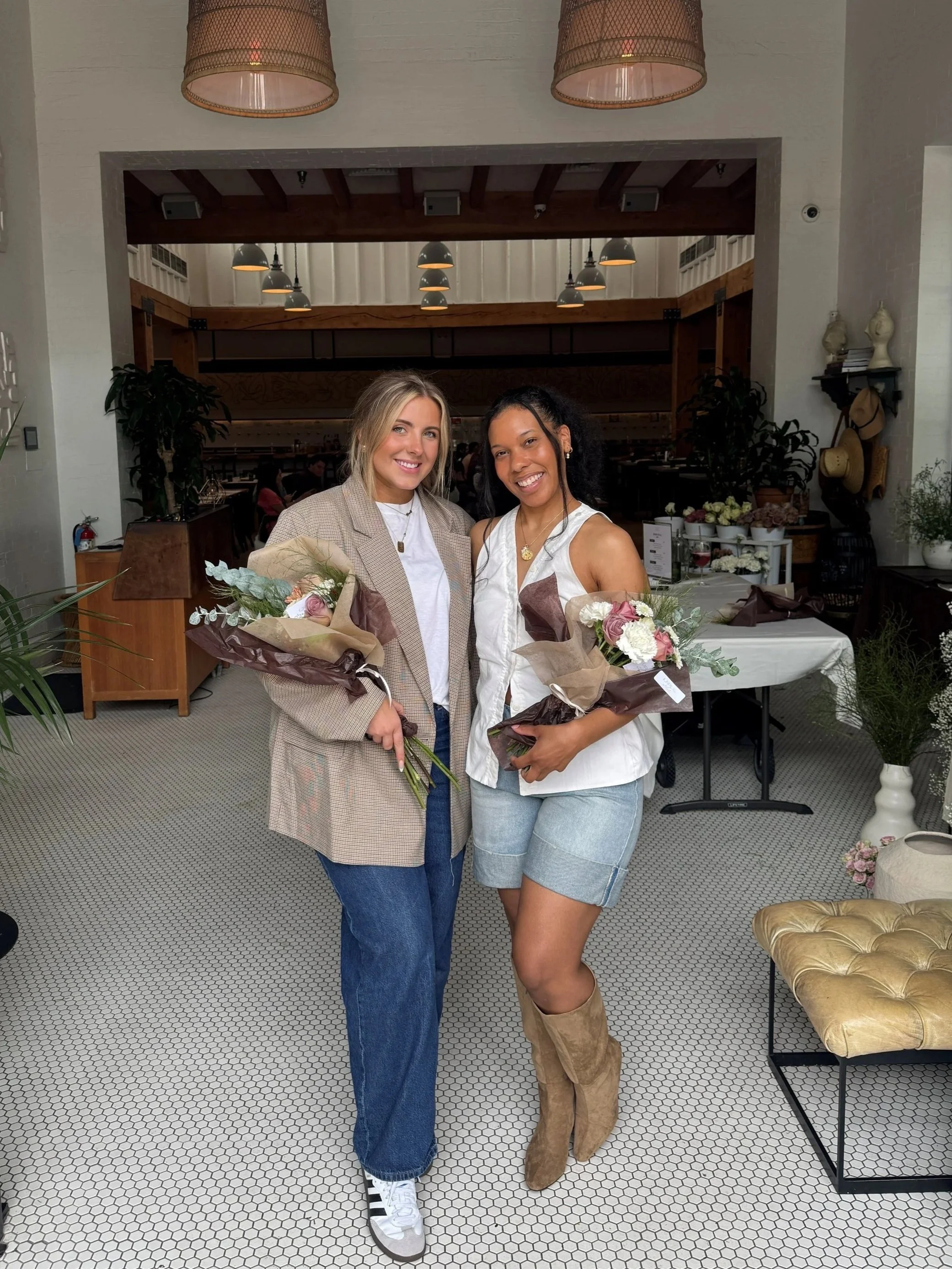 Two women smiling and holding bouquets of flowers standing indoors in a decorated space.