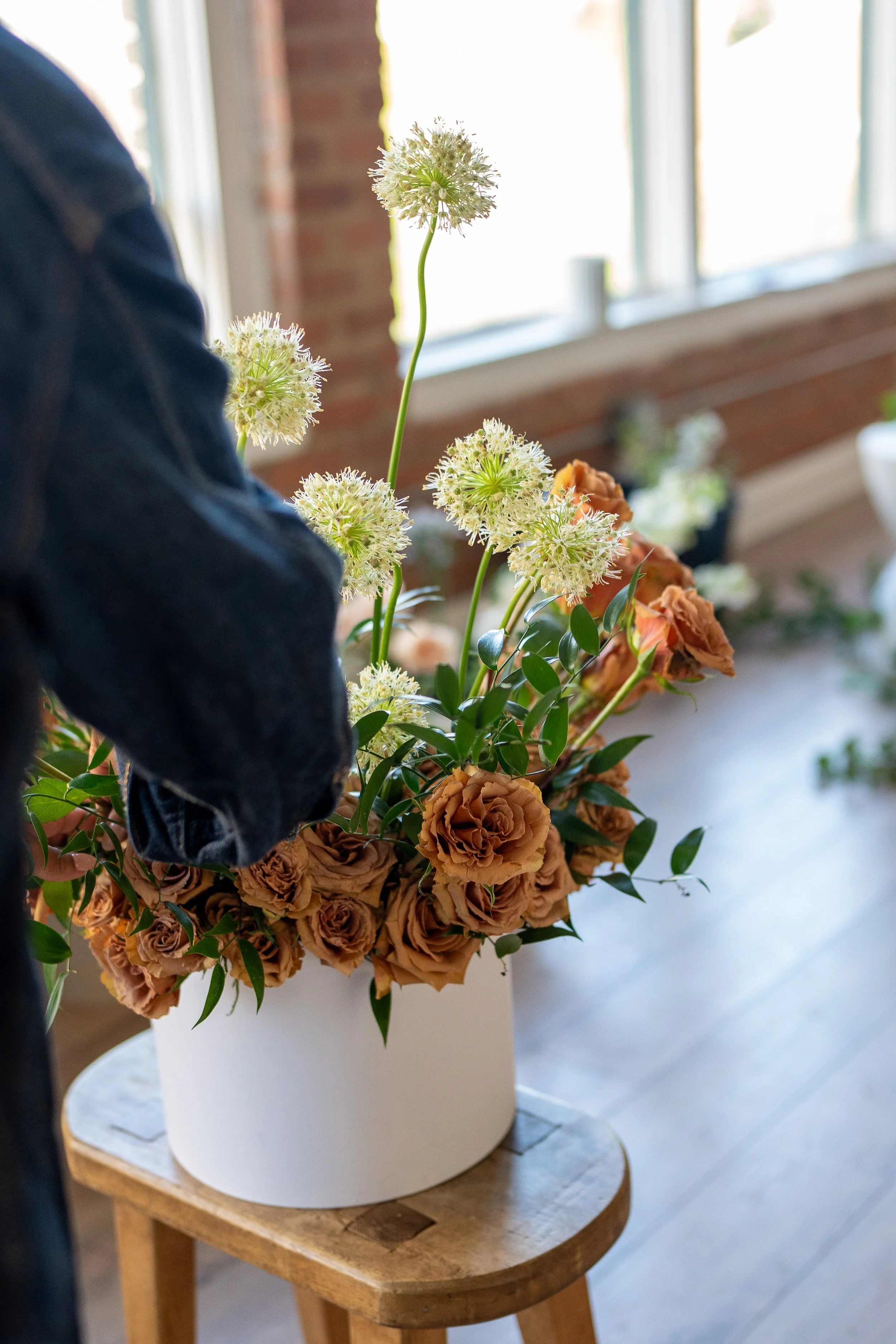 Person arranging a large bouquet of flowers with brown roses and white globe-like flowers in a white cylindrical vase, placed on a wooden stool near a window.