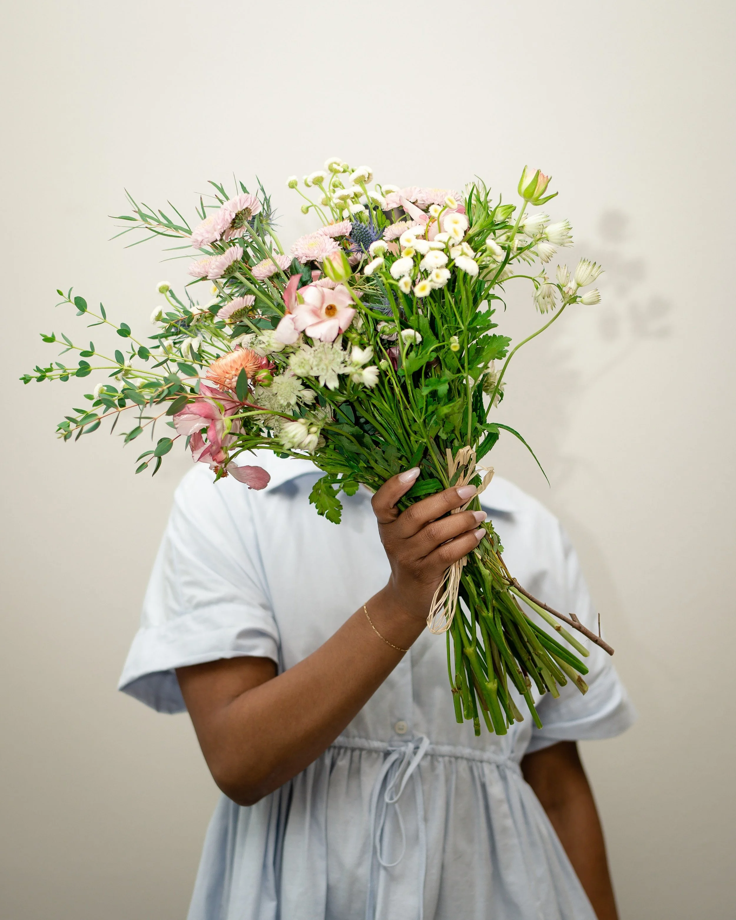 A person holding a large bouquet of mixed colorful flowers in front of their face, wearing a white buttoned dress with puffed sleeves.