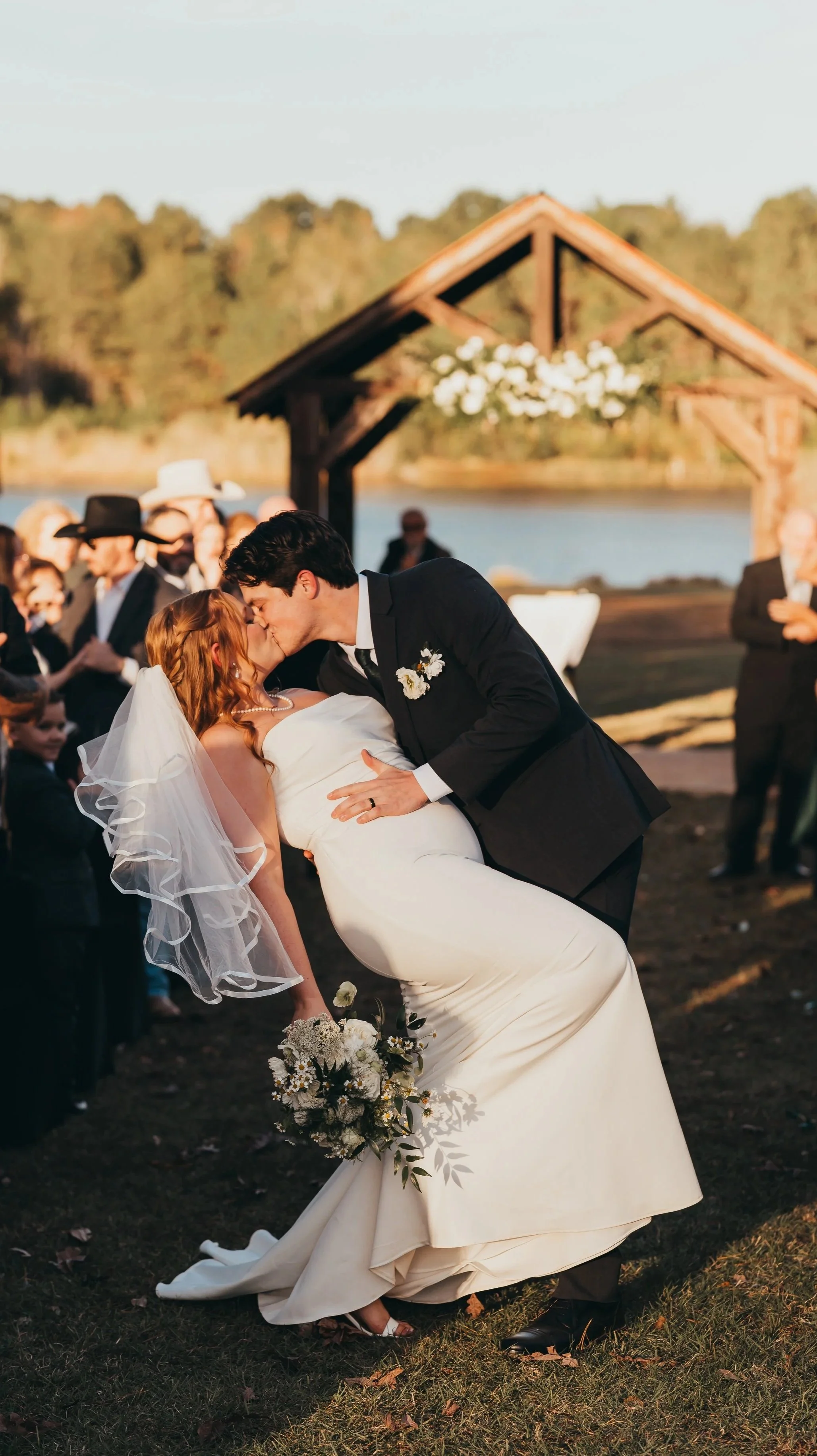 A newly married couple sharing a kiss during an outdoor wedding ceremony, show casing her bouquet, his boutonnière, and the floral installment above the alter.