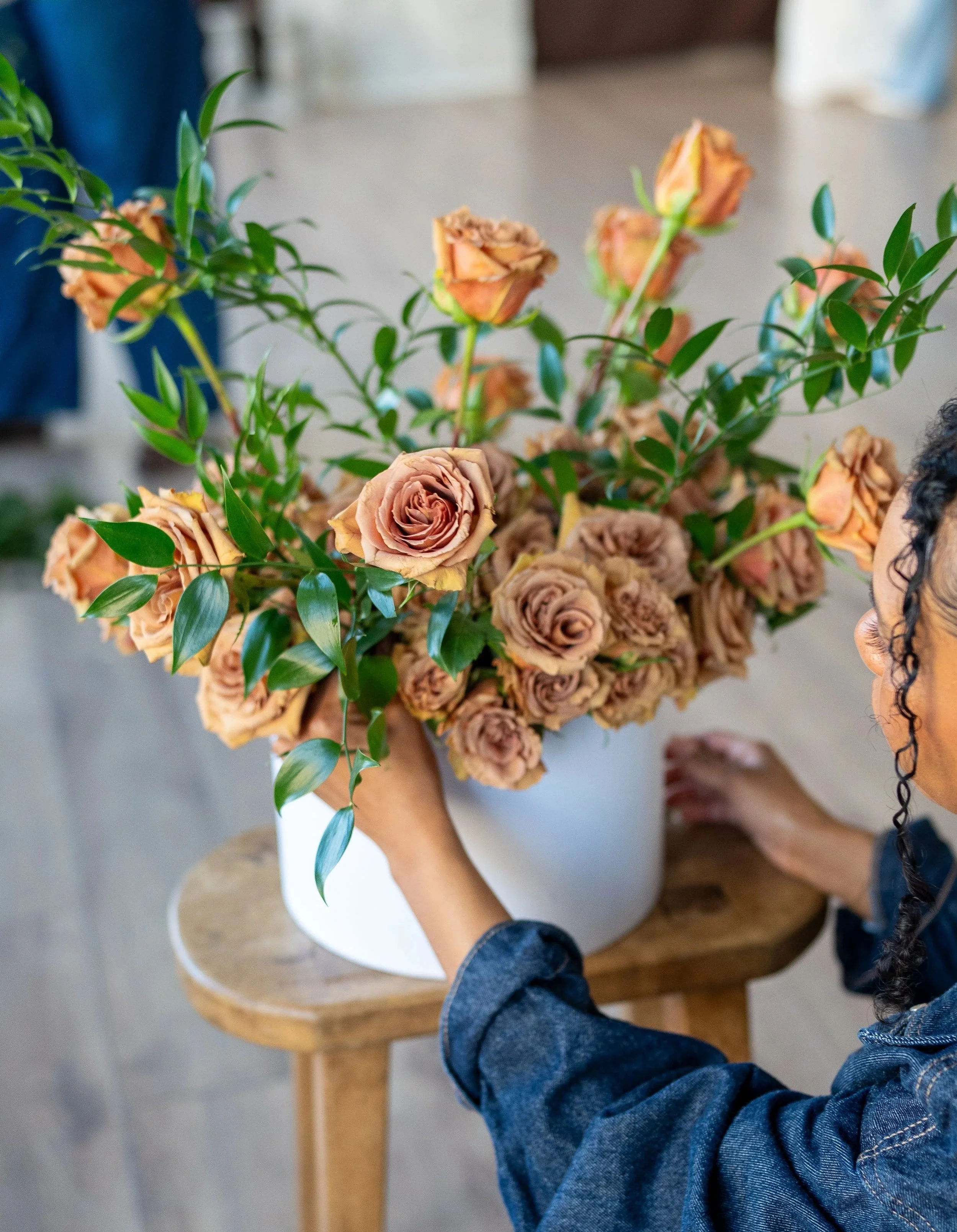 A person arranging a bouquet of brown-colored roses with green leaves in a white vase placed on a wooden stool.