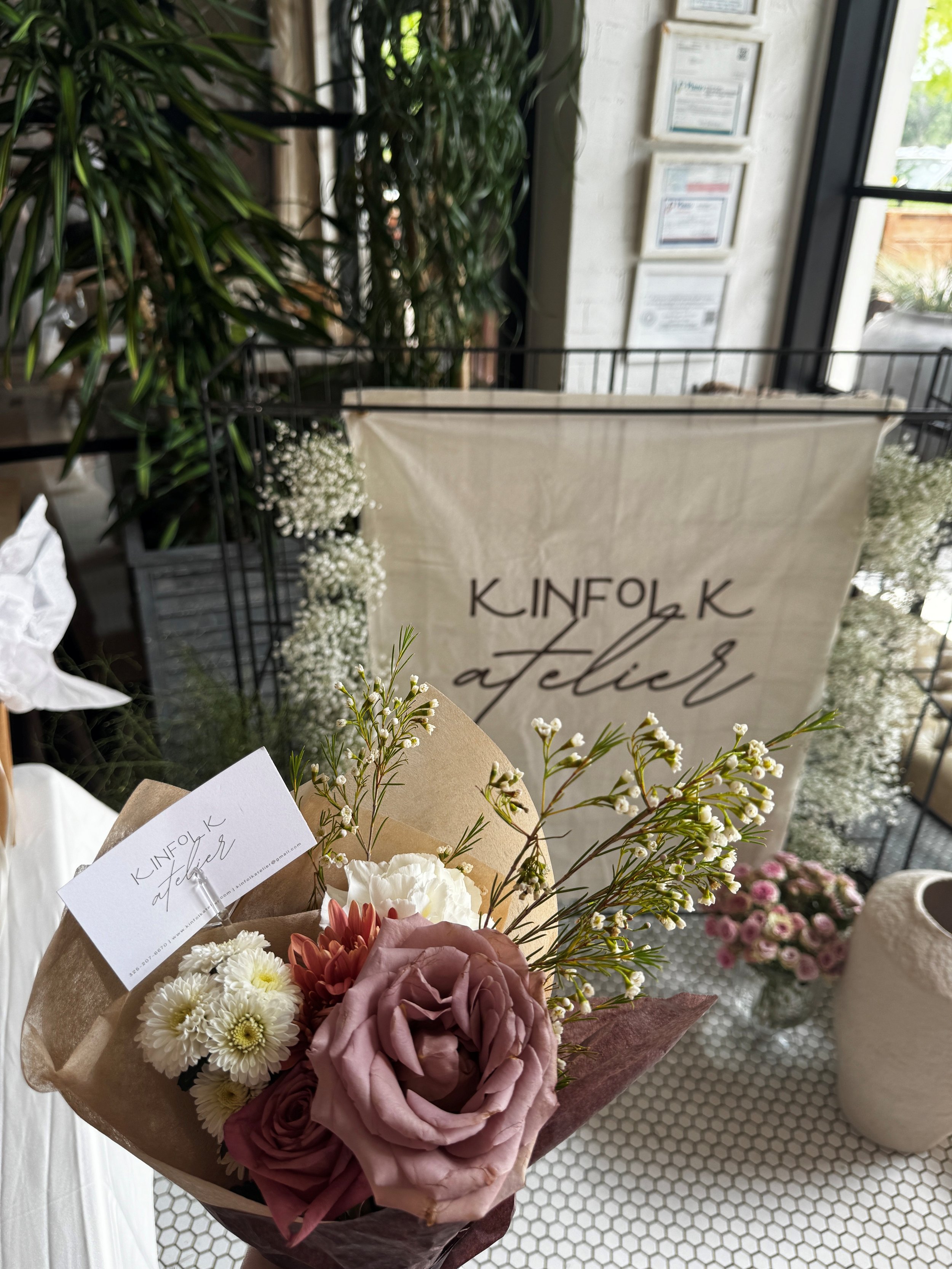 A bouquet of pink, white, and purple flowers with greenery, wrapped in brown paper, in front of a display with a sign that reads 'KINFOLK atelier', inside a floral shop with large plants.