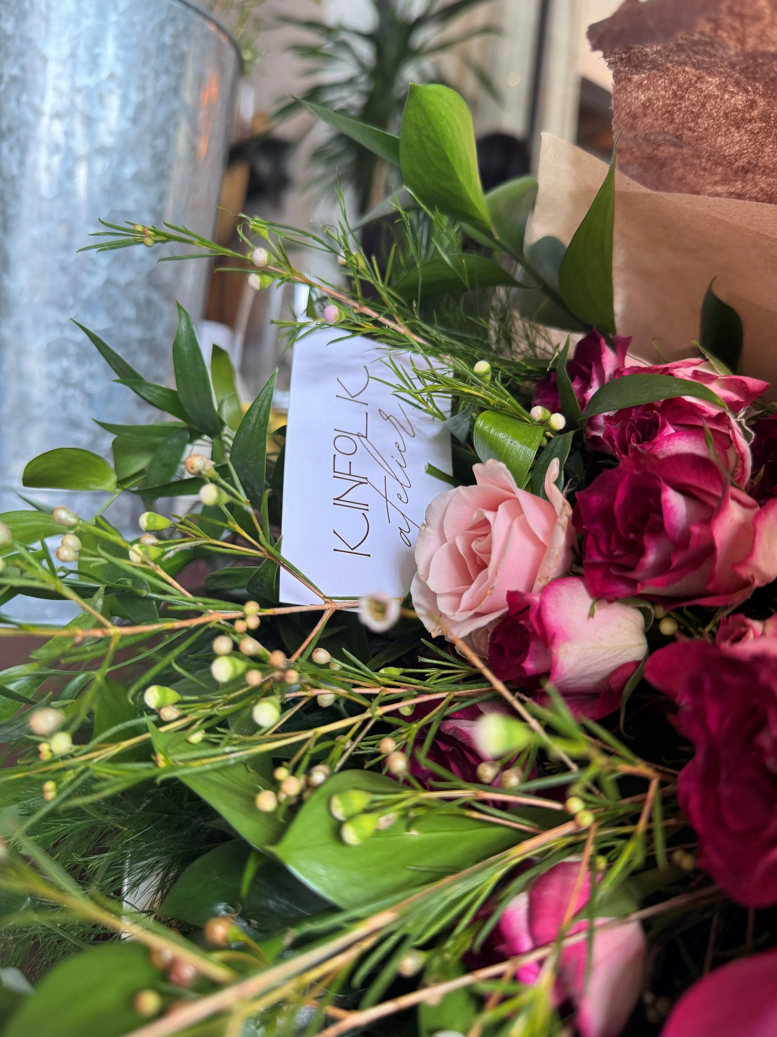 Close-up of a floral bouquet with pink and dark pink roses, green leaves, and white berry-like accents, with a note attached that reads "KINFOLK atelier."