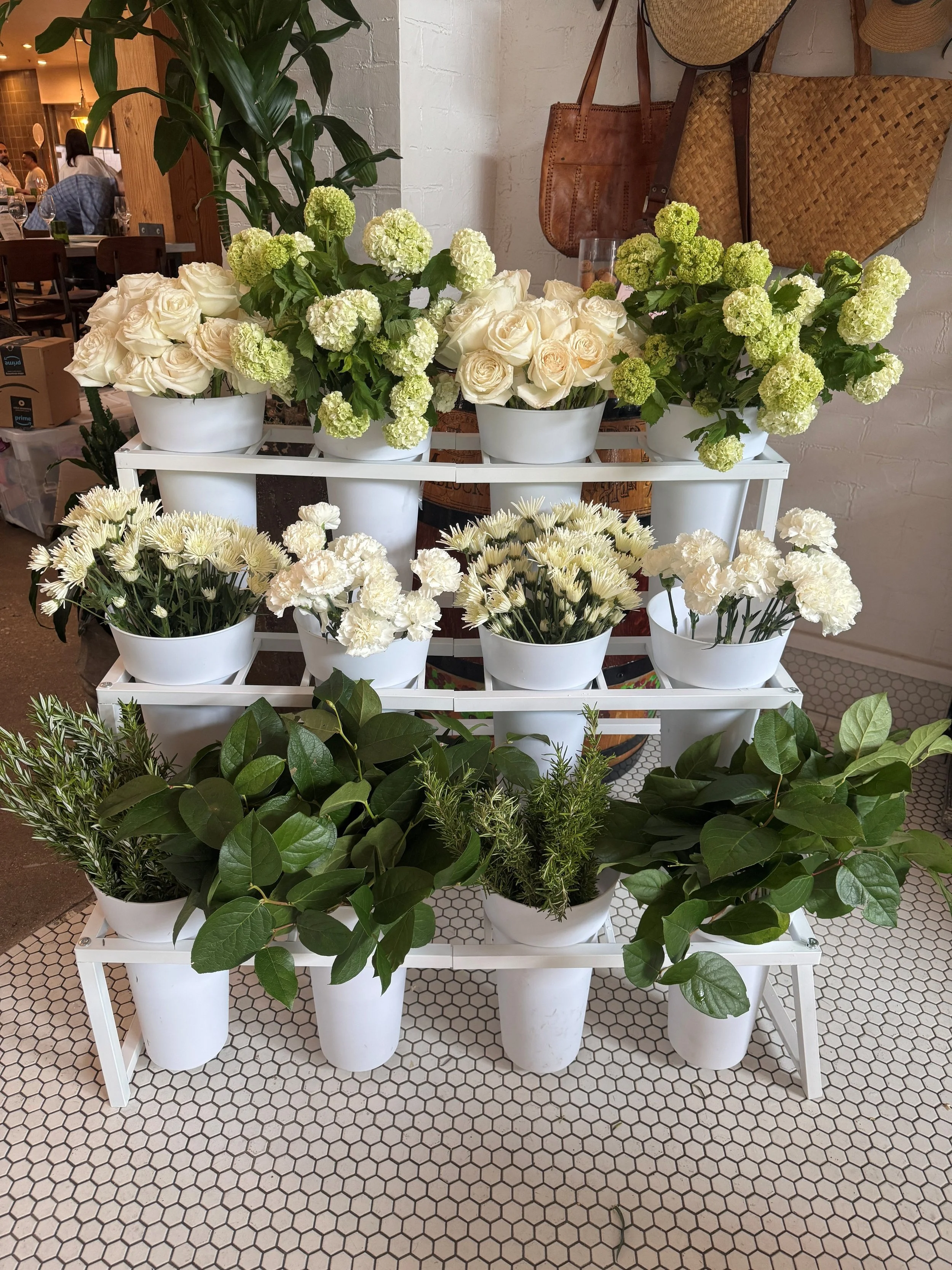 A display of white roses, hydrangeas, carnations, daisies, and various green plants in white pots on a white stand inside a café.