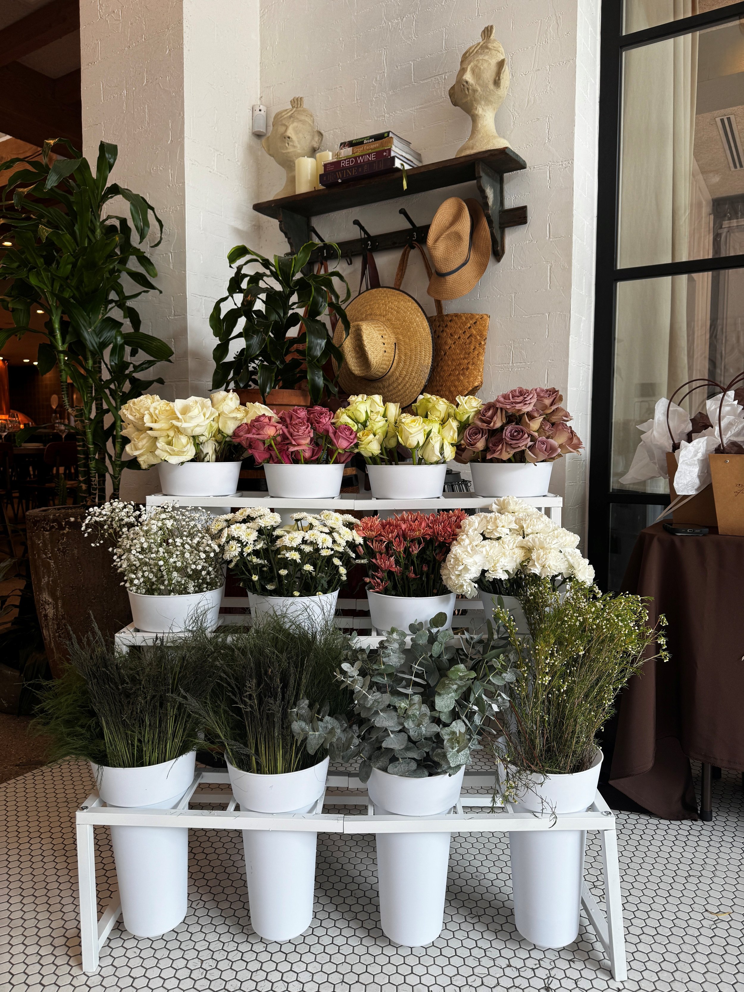 Display of various flowers  such as roses and other greenery in white pots on a white multi-tiered stand, with hats hanging on a wall above and a brick wall in the background.