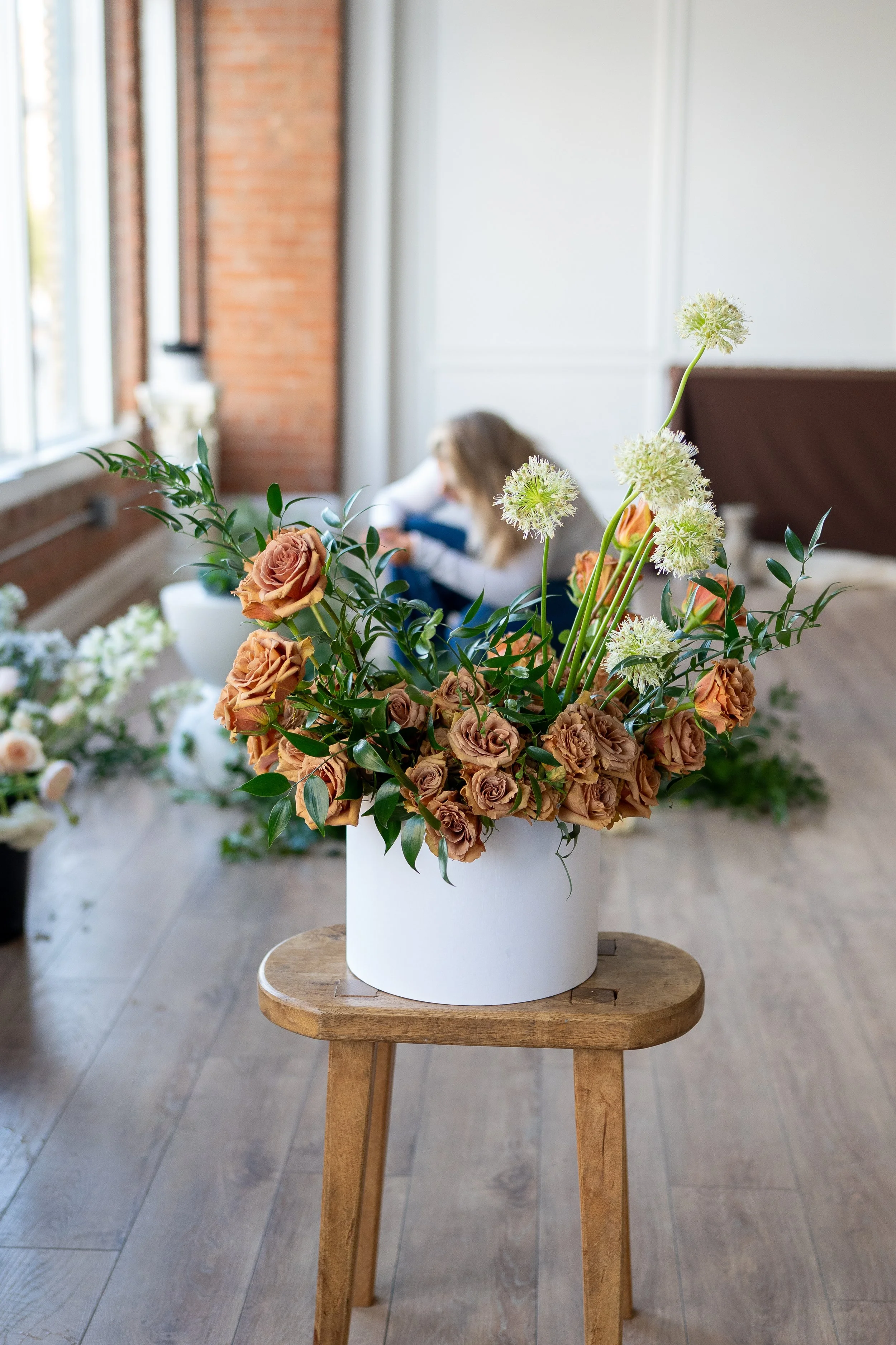 A white round flower vase containing brown roses and white spherical flowers sits on a small wooden stool in a bright room with large windows.