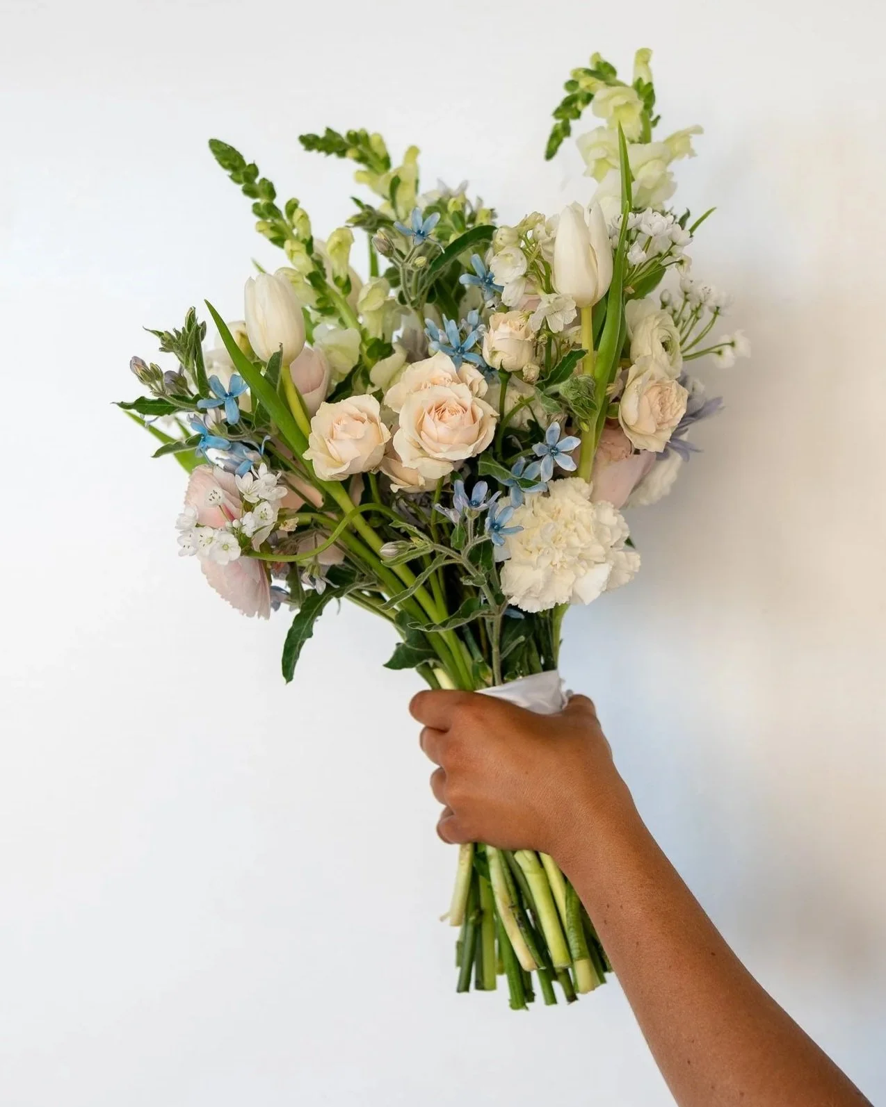 Person holding a wedding bouquet of mixed white and pastel flowers, including roses and tulips, along with green foliage to fill out the bouquet.
