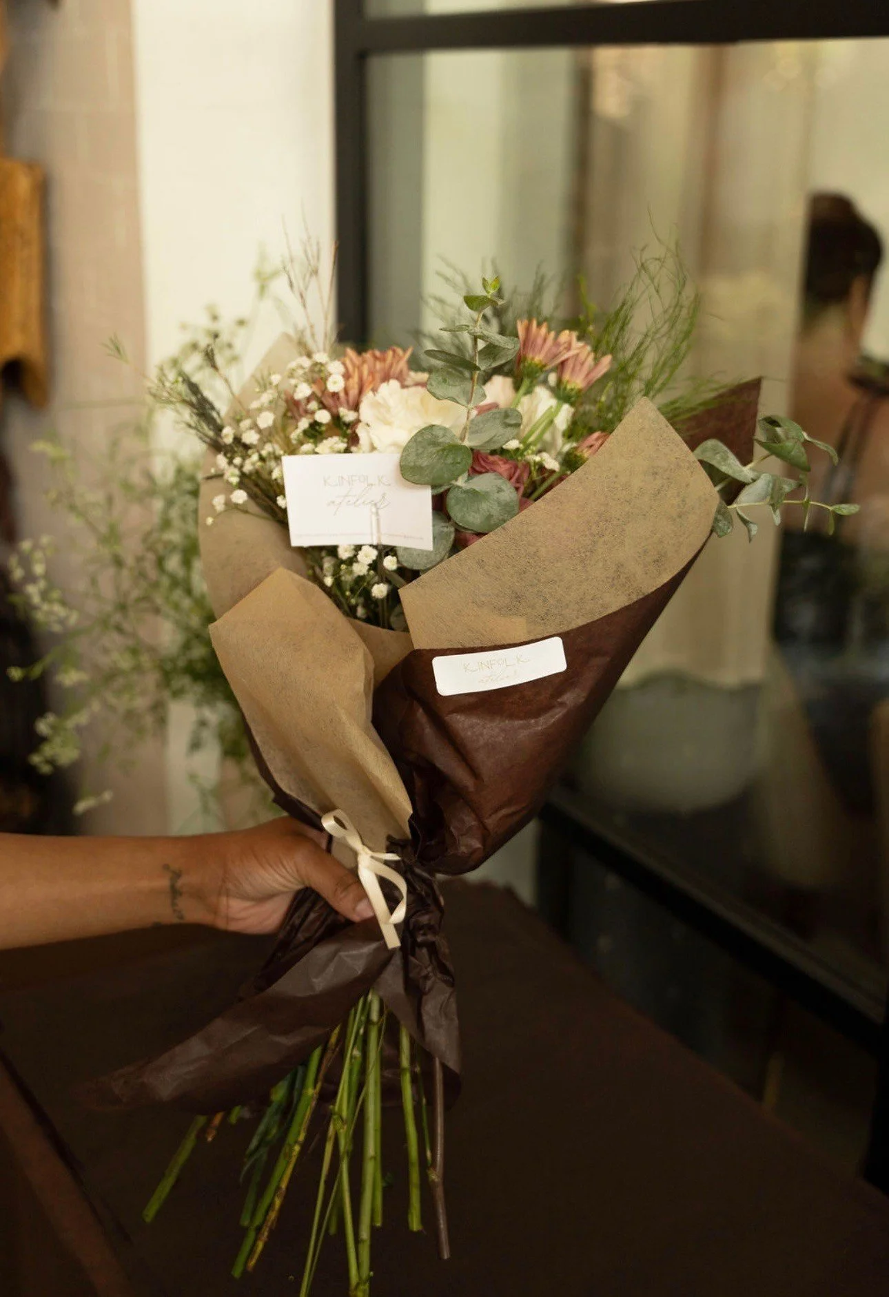 A bouquet of mixed flowers including pink, white, and green foliage, wrapped in brown and beige paper, being held by a person's hand.