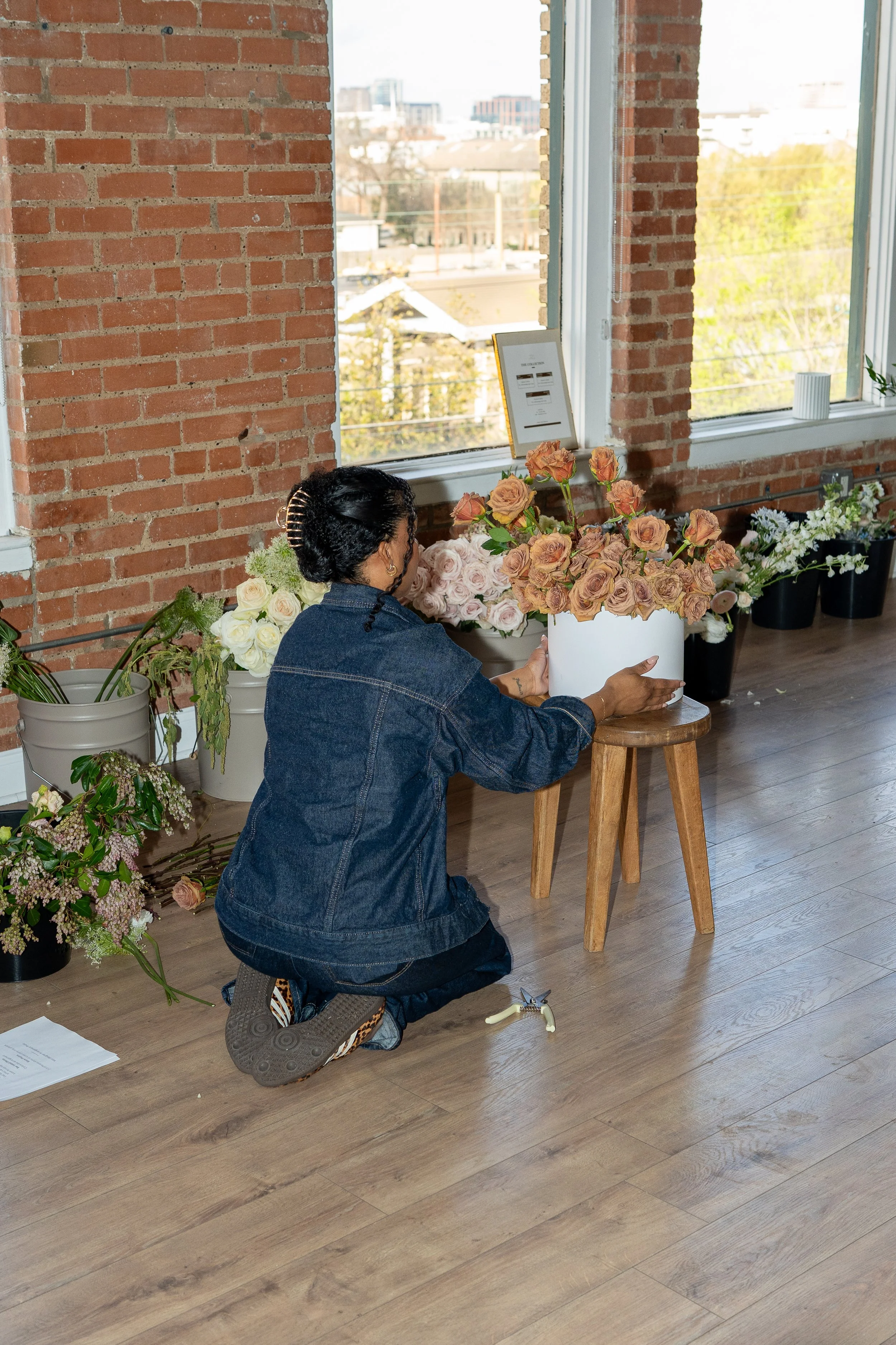 A woman arranging brown and pink roses in a white vase inside a room with large windows and a brick wall.