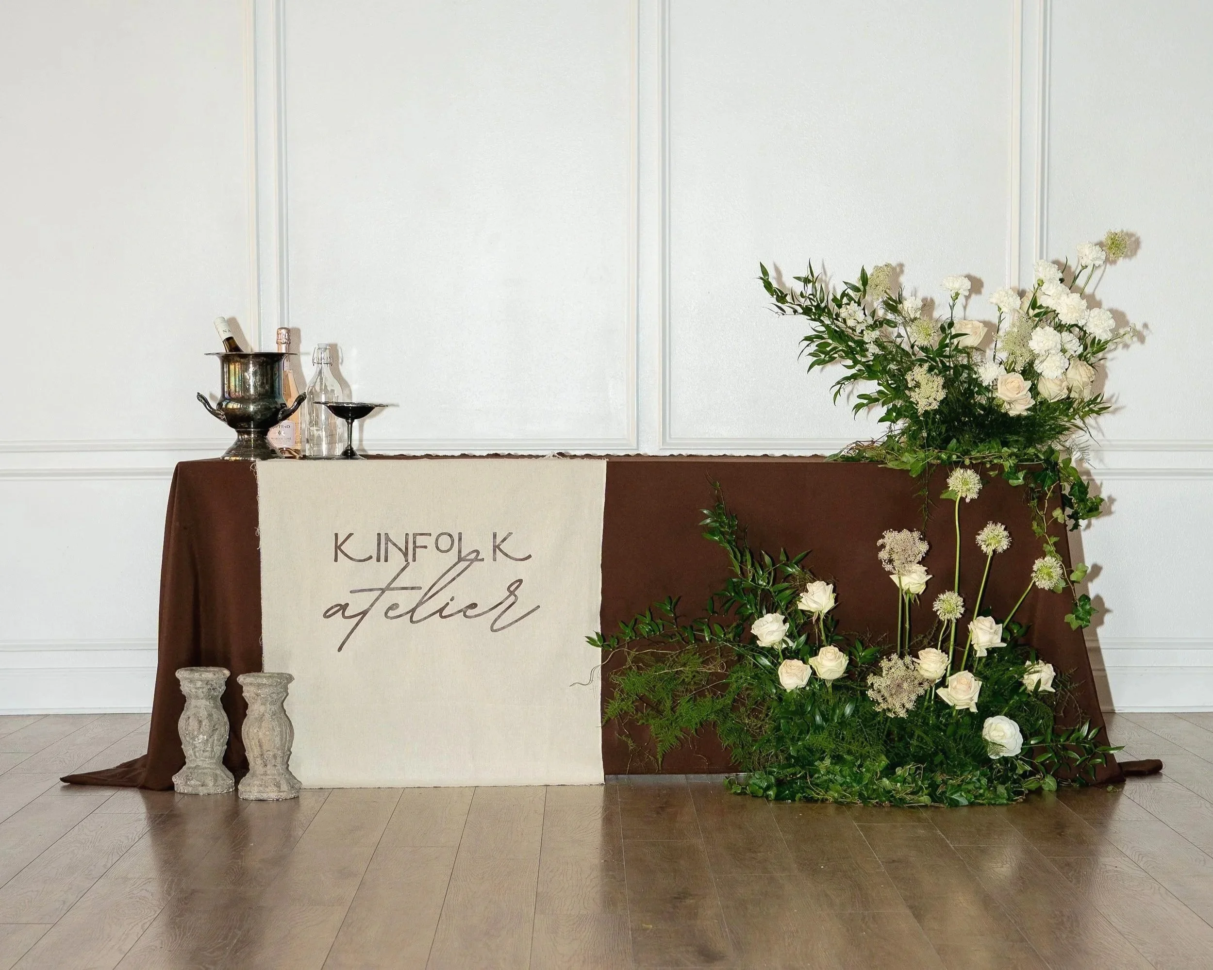 A decorated table with a carefully curated wedding or event floral installment, adorned with white roses, white and green foliage, and a large flower arrangement.