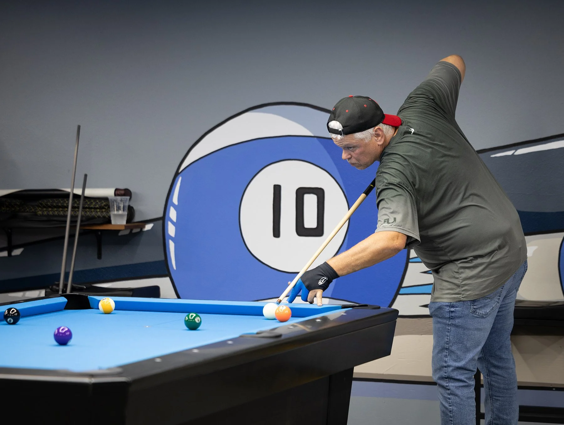 A man playing pool on a pool table in a room decorated with a large Frank Sinatra-themed mural with a clock showing 10.00. The man is wearing a cap backwards, gray T-shirt, and blue jeans, and is aiming to take a shot.